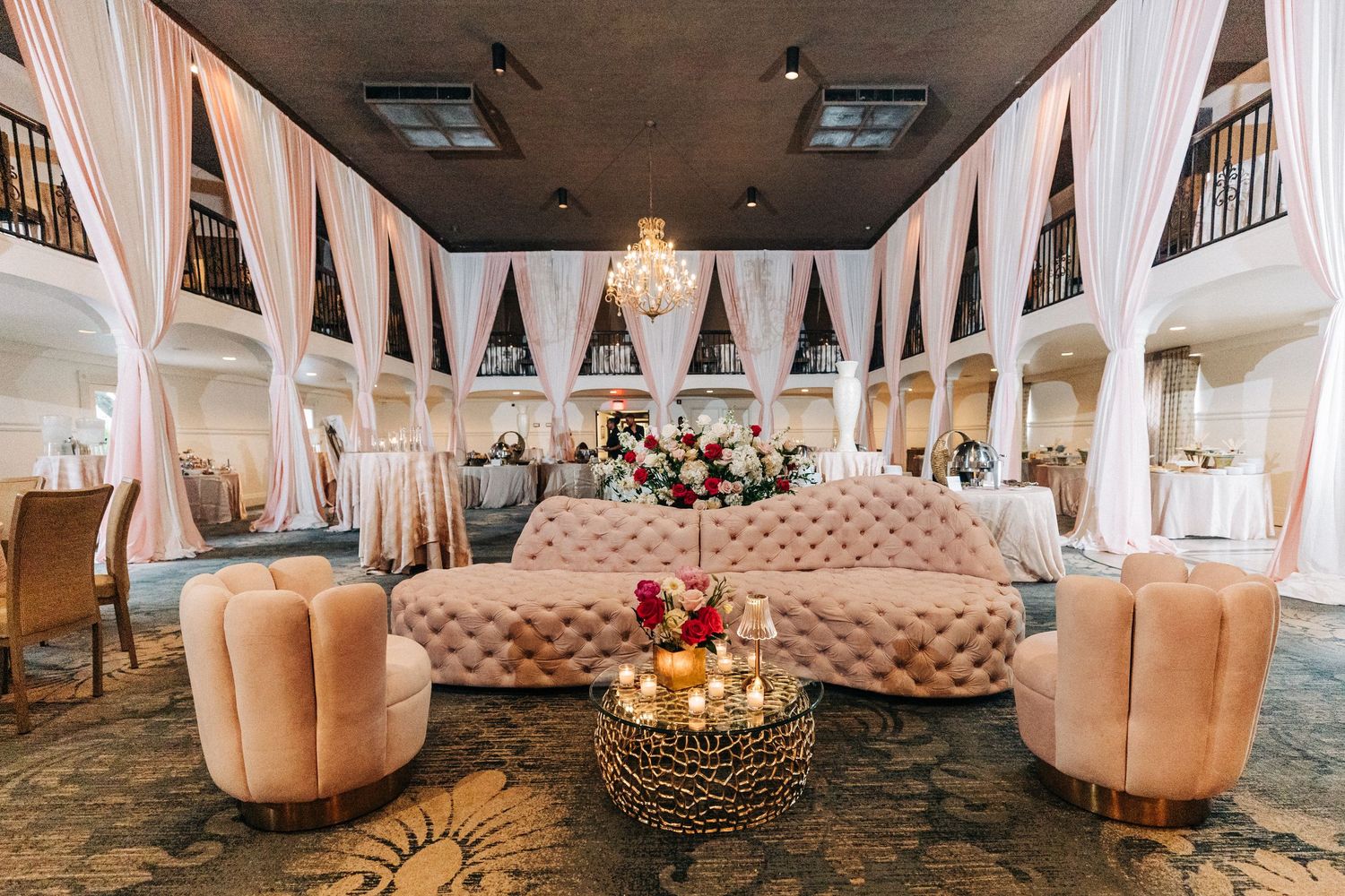 An elegant wedding reception venue with white draping, chandeliers, and plush velvet seating in blush and cream tones.