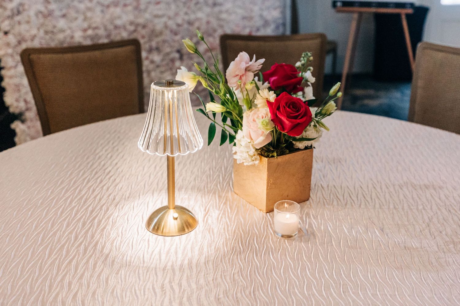 Small gold table lamp and wooden box with fresh flowers on white tablecloth covered round table.