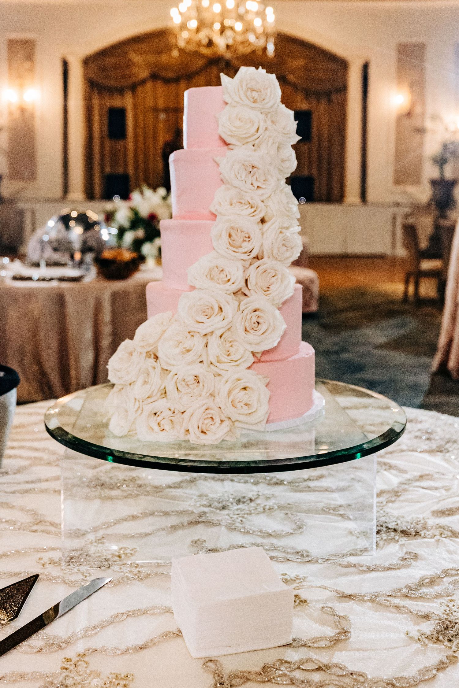 A five-tier pink ombre wedding cake decorated with cascading white roses on a glass stand.