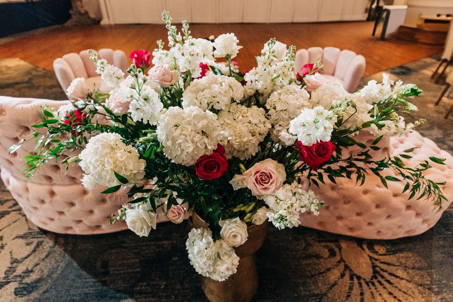 A wedding ballroom decorated with elegant floral arrangements featuring white hydrangeas, roses and greenery on pink chairs.