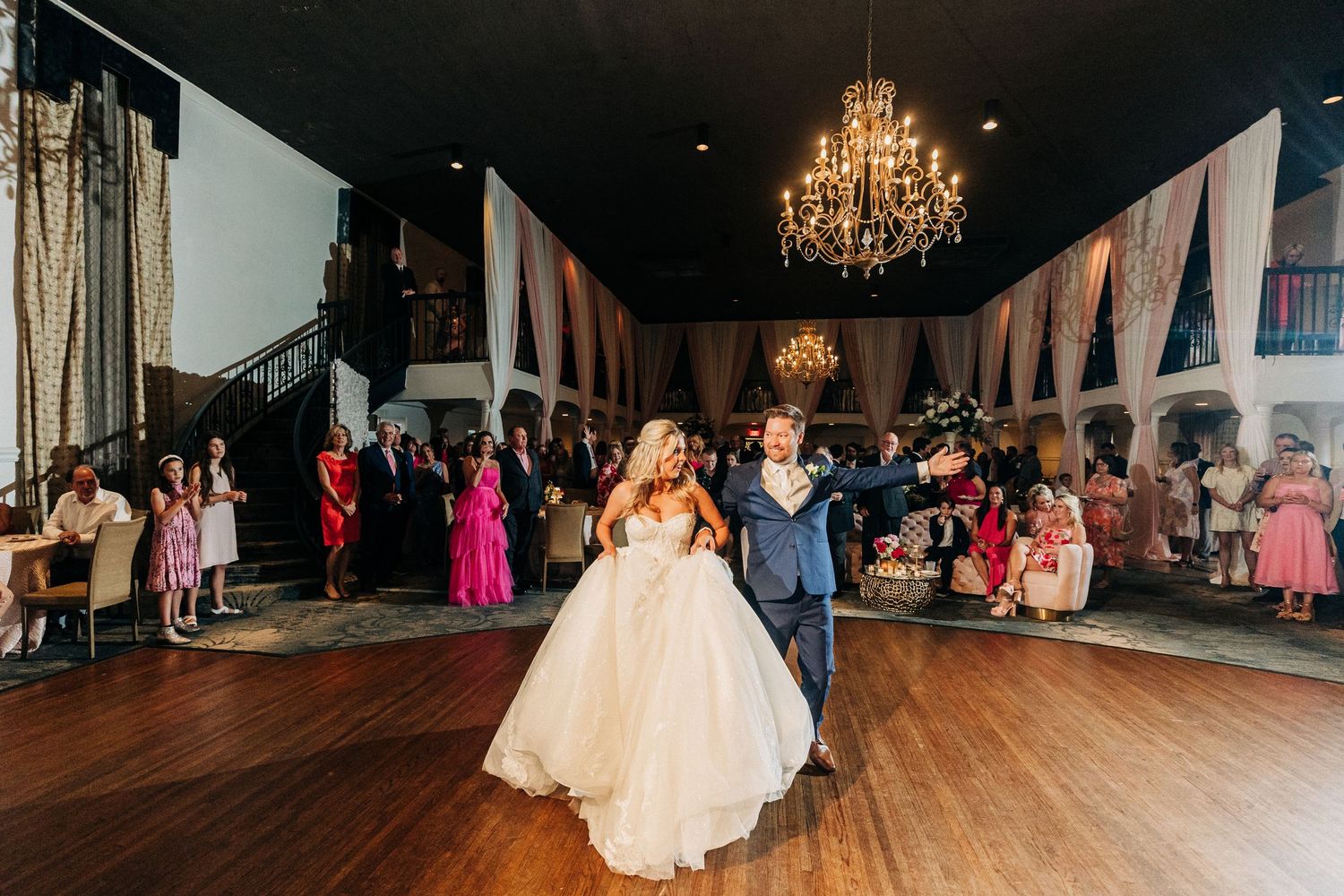 Elegant ballroom wedding reception venue with chandeliers and guests dancing on dark hardwood floors.