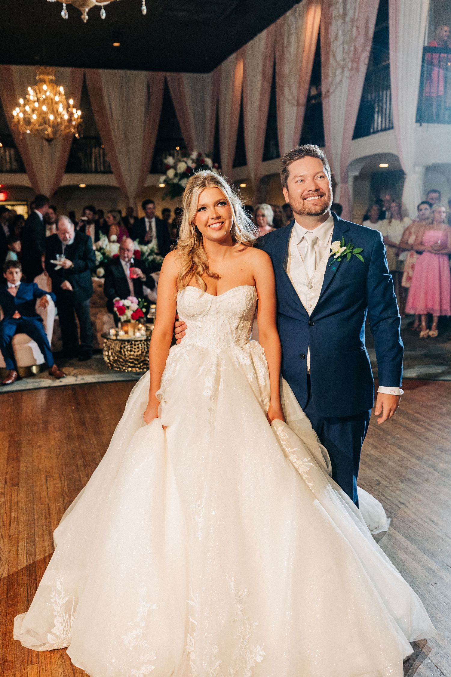 A bride in a strapless ballgown and groom in navy suit stand together at their wedding reception under crystal chandeliers.