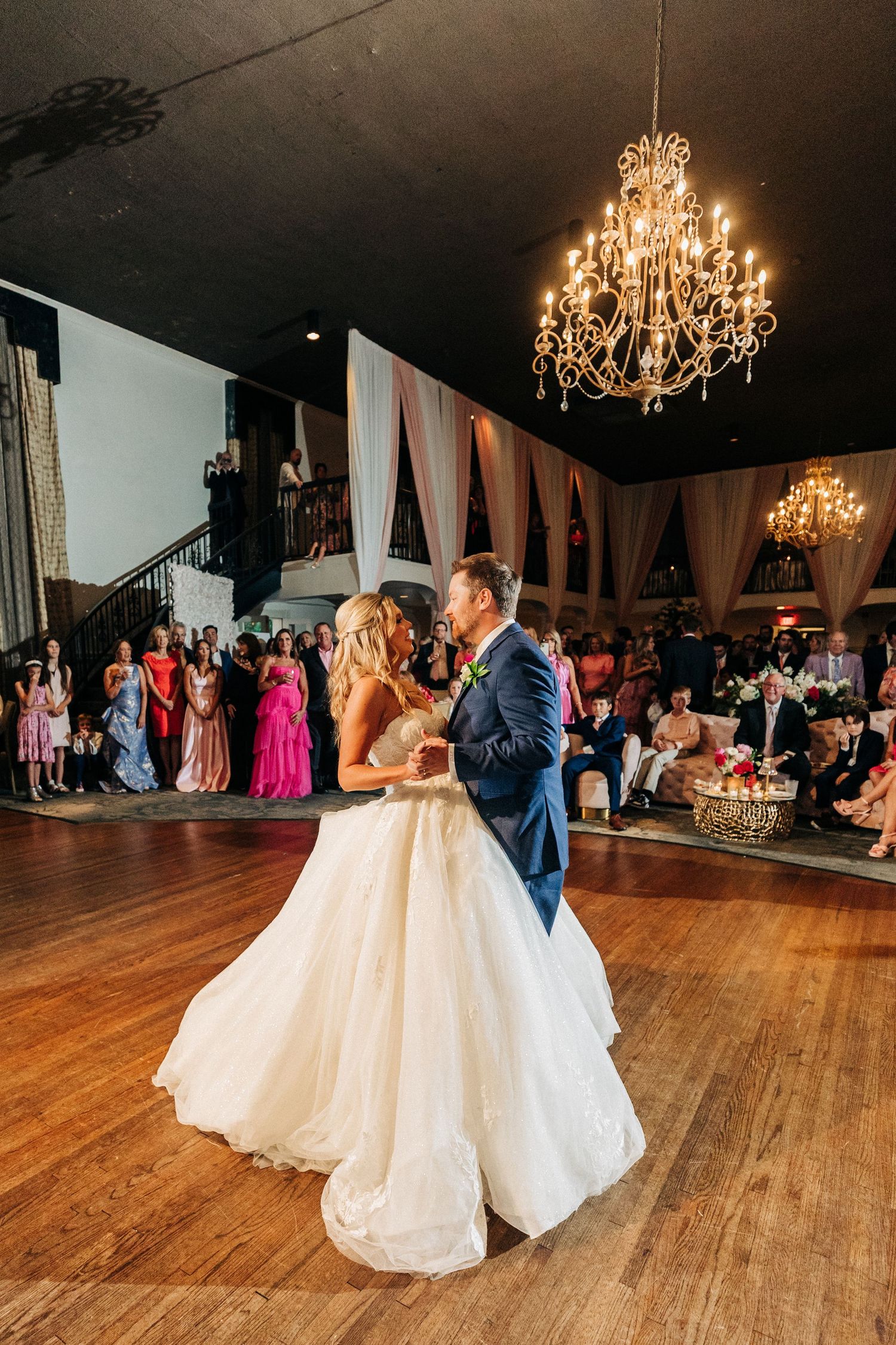 A couple shares their first dance on a hardwood floor at their wedding reception with chandeliers overhead.
