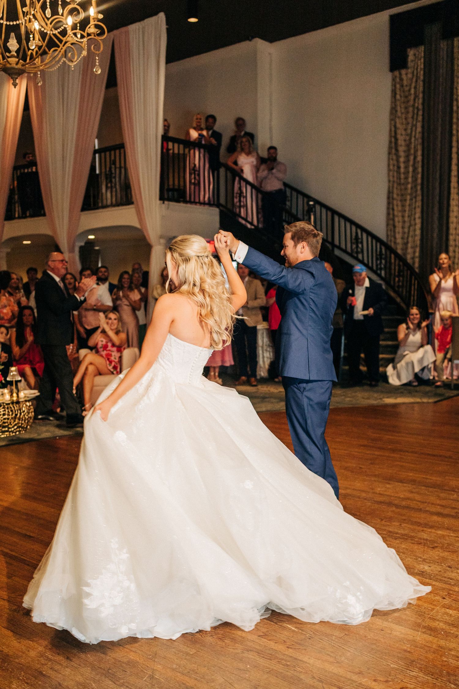 Couple performs their first dance at an elegant wedding reception with guests watching from a curving staircase.