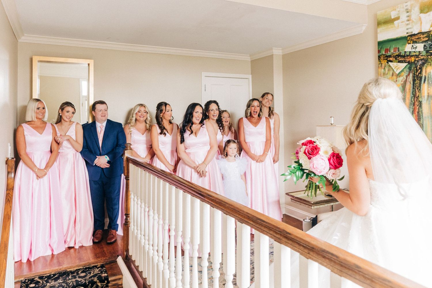 Wedding party wearing pink dresses and a navy suit gather on staircase to watch person in white dress.