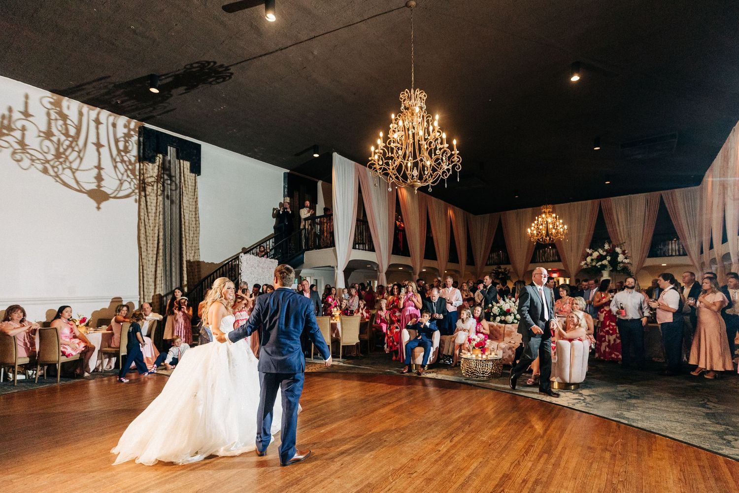 A couple performs their first dance in a ballroom decorated with elegant chandeliers and curtains as guests watch.