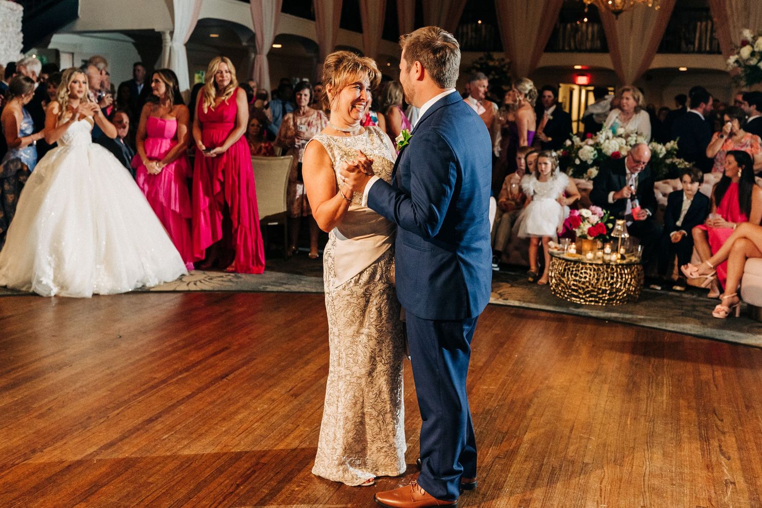 A couple shares an intimate dance moment on the dance floor at a wedding reception with bridesmaids in red dresses looking on.