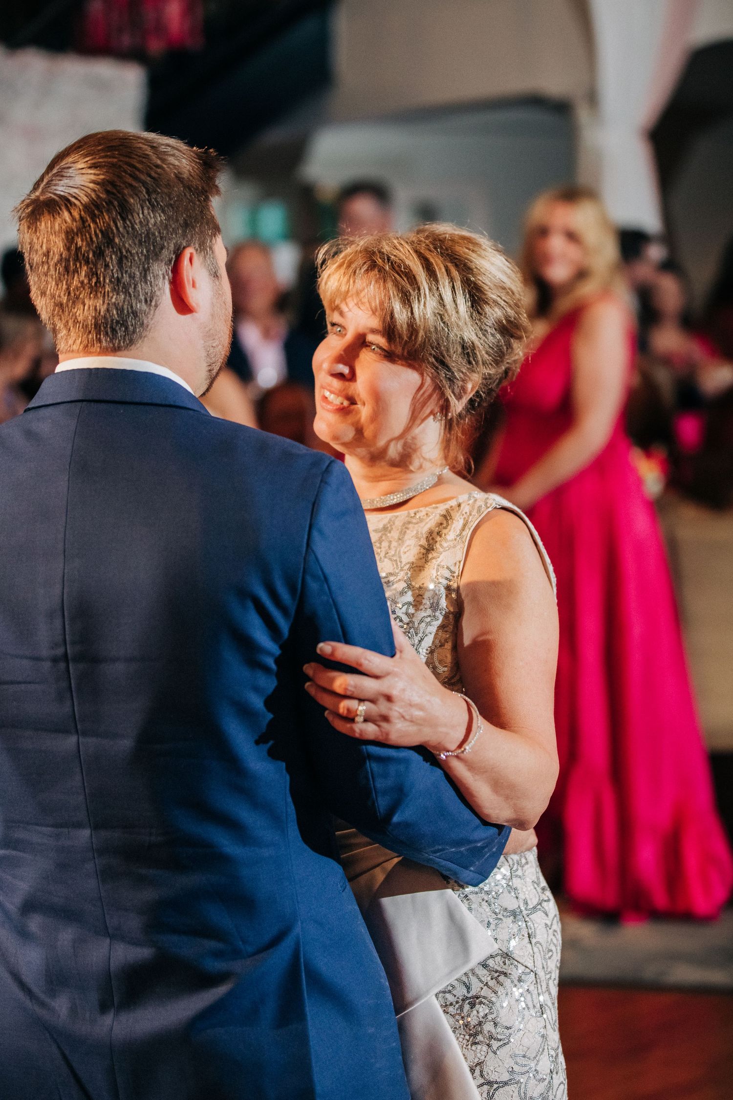A couple shares an intimate dance moment at a wedding reception, with a guest in a bright pink dress visible in background.