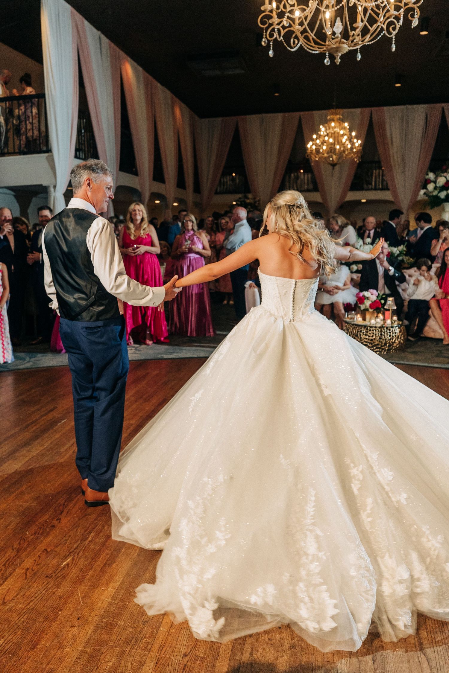 A bride in a princess ballgown spins on the dance floor with someone in a vest during a reception lit by chandeliers.