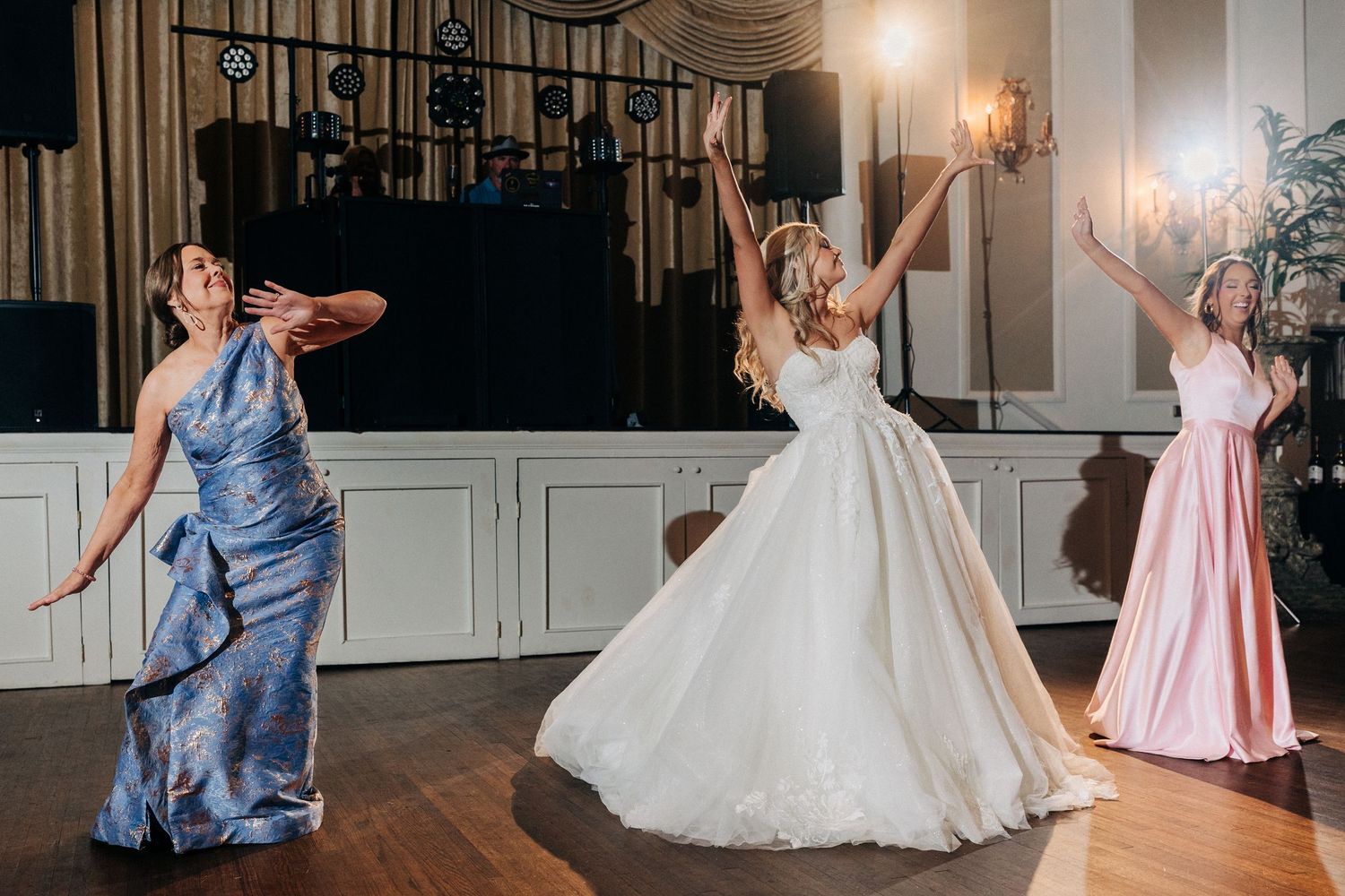 People dancing joyfully on a wooden floor at a wedding reception with elegant wall sconces in the background.