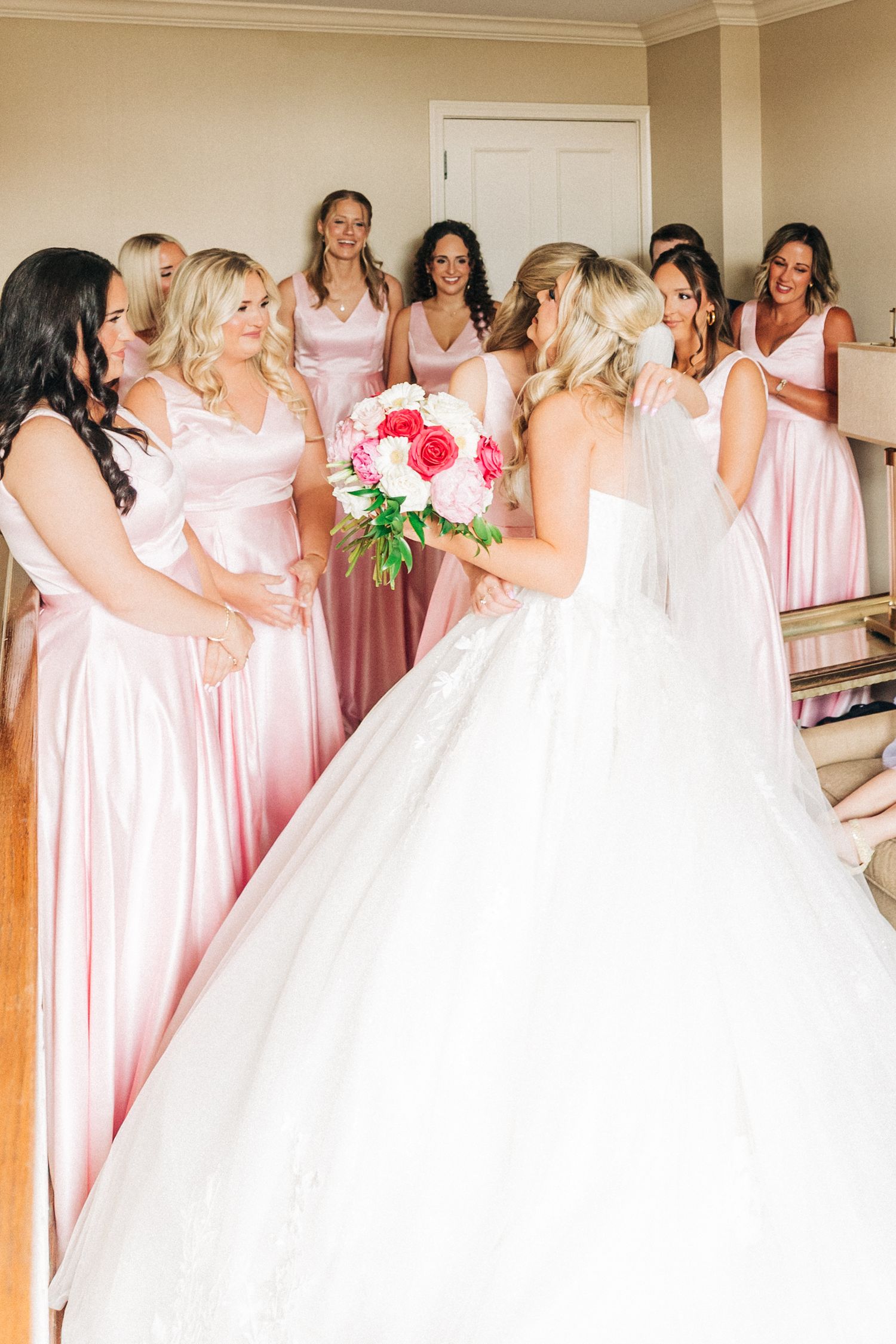 Bridesmaids in pink dresses embrace and celebrate with the bride in a white wedding gown during a getting ready moment.
