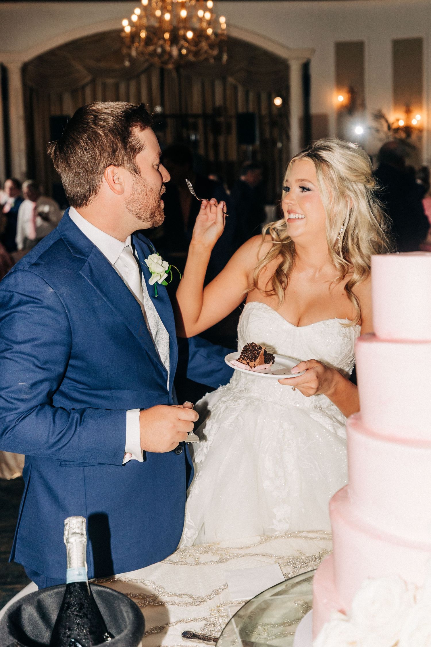 Newlywed couple playfully feeds each other wedding cake at their reception under elegant chandelier lighting.