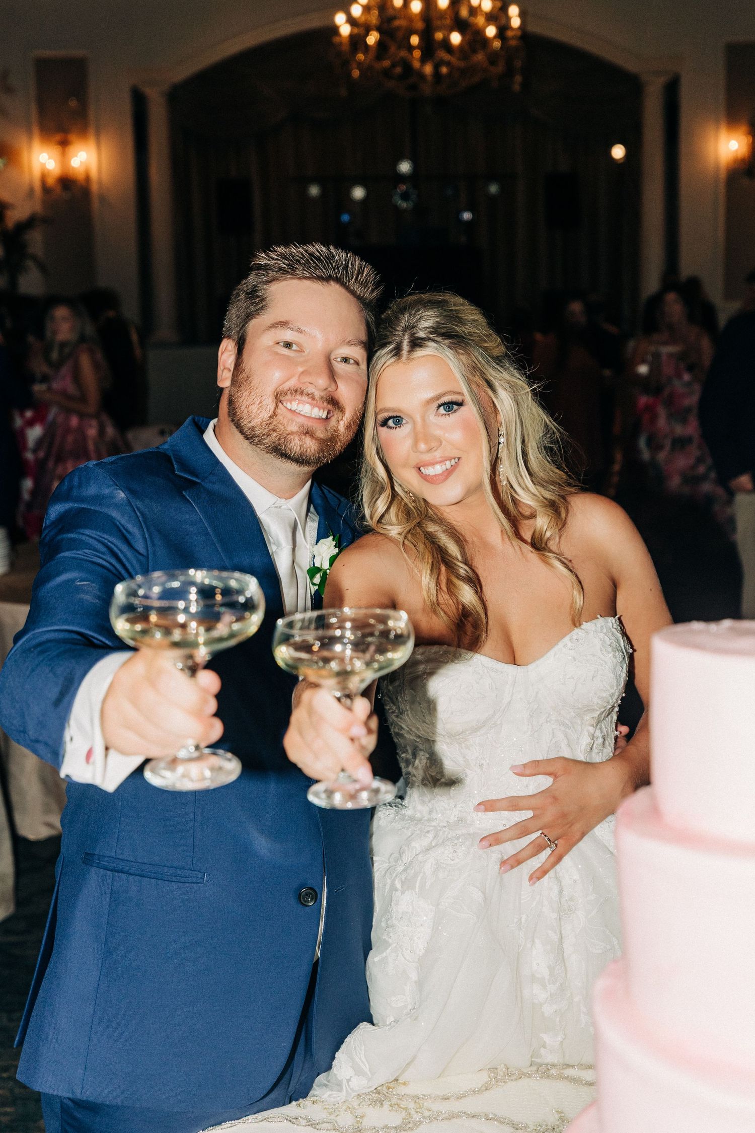 A newlywed couple toasts with wine glasses at their wedding reception beside a pink wedding cake.
