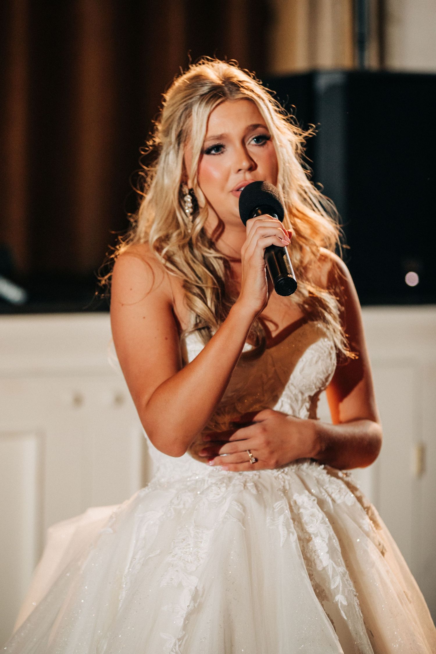 A bride in a white lace wedding dress sings into a microphone during her wedding reception.