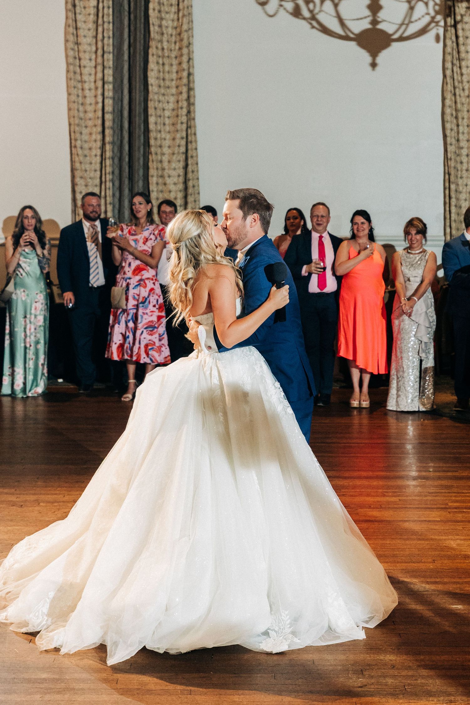 A couple shares their first dance at their wedding reception on a hardwood floor while guests watch from the background.