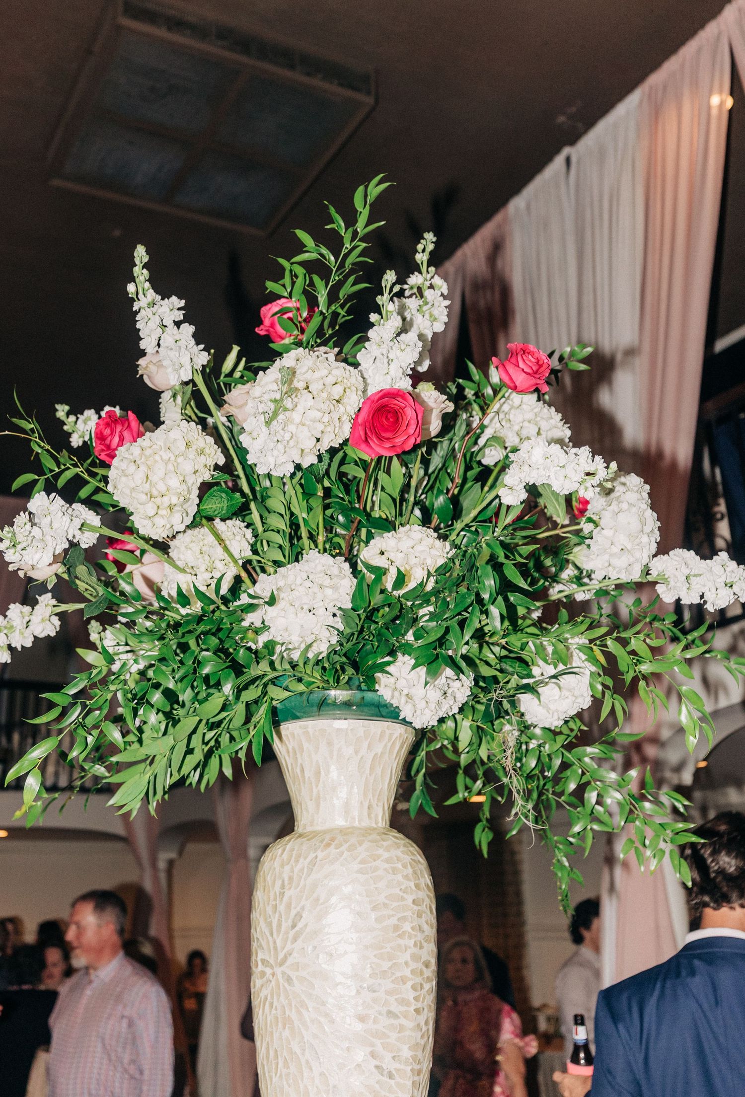 Large floral arrangement with white hydrangeas, pink roses and greenery in a textured white vase at an indoor event.