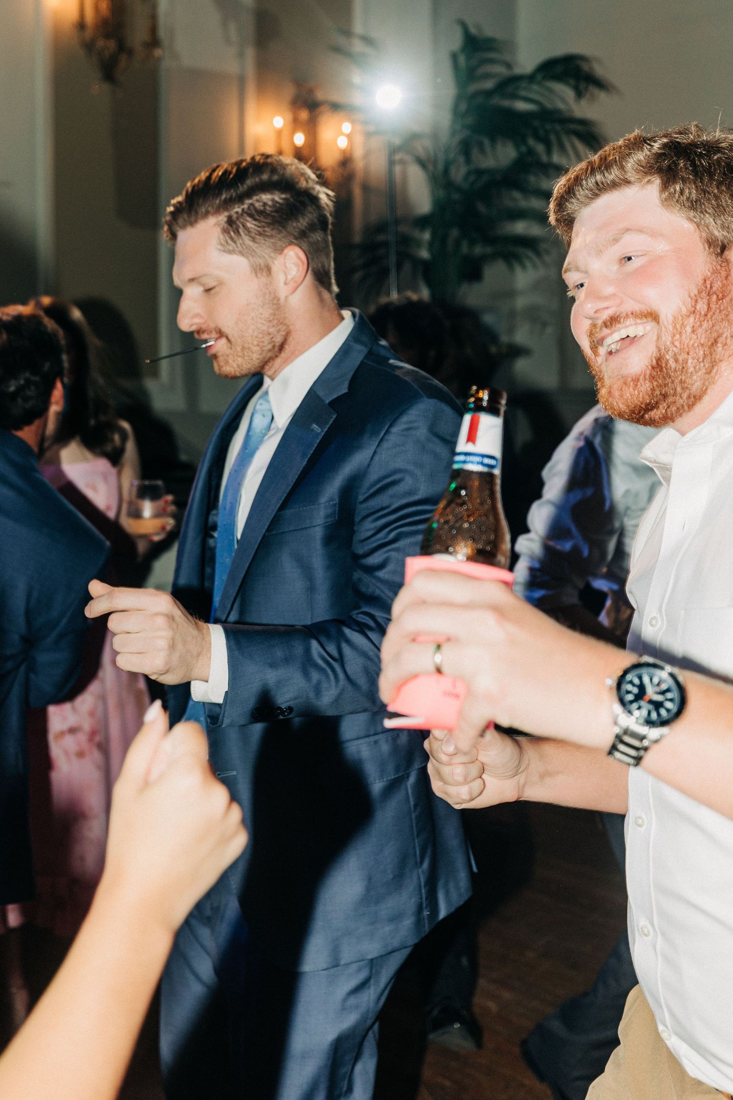 Party guests dancing and enjoying drinks at an indoor celebration with warm lighting and decorative palm plants.