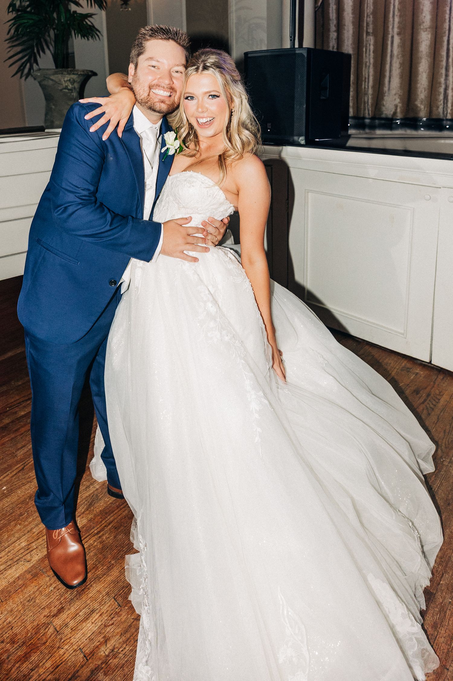A joyful couple shares a dance at their wedding reception on a wooden floor in a light-filled venue.