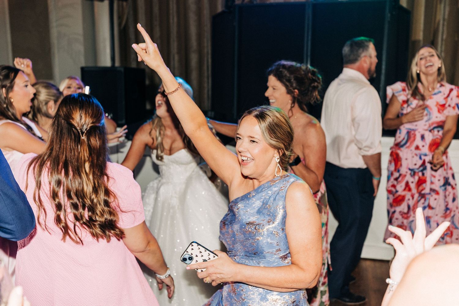 A large group of people dance and celebrate at an event, with colorful outfits visible in the dimly lit space.
