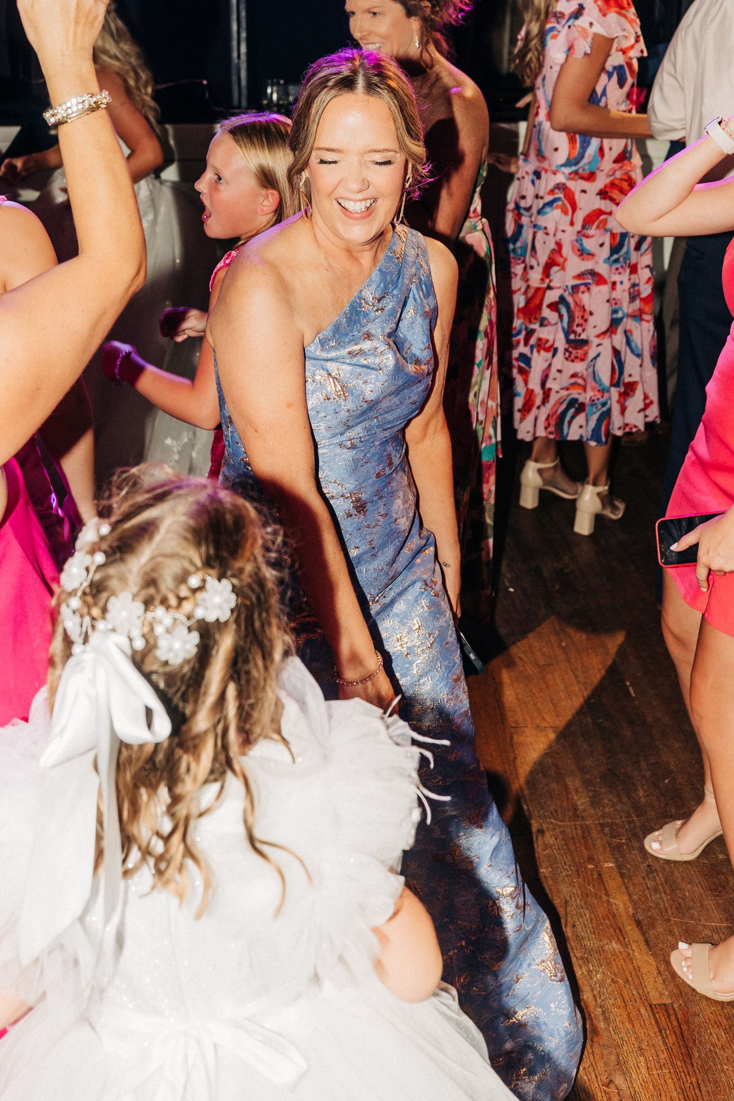 A lively dance party scene on a wooden floor with people dancing in colorful dresses and casual attire.