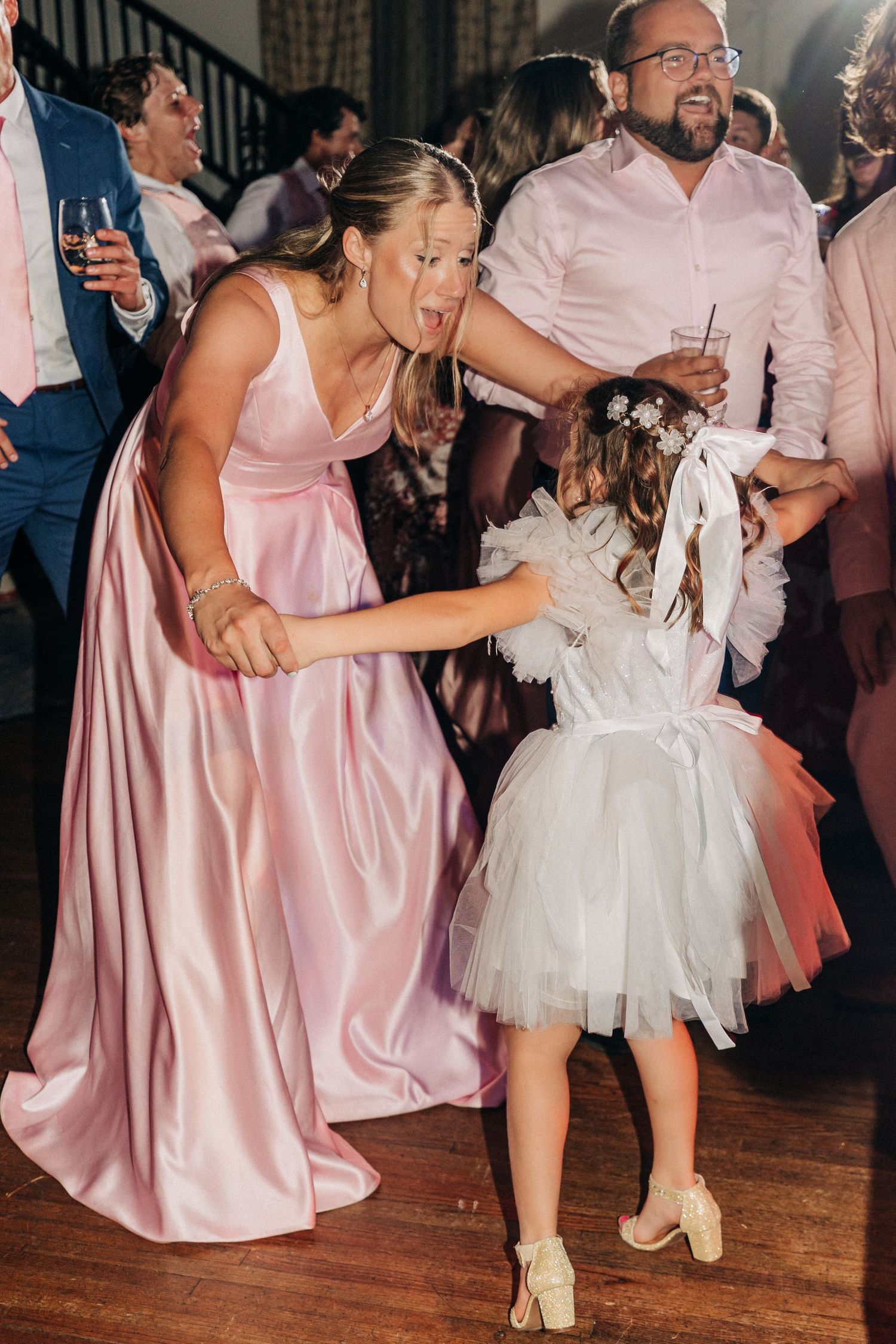 A bridesmaid in a pink satin gown dances with a young flower girl in white tulle at a wedding reception.