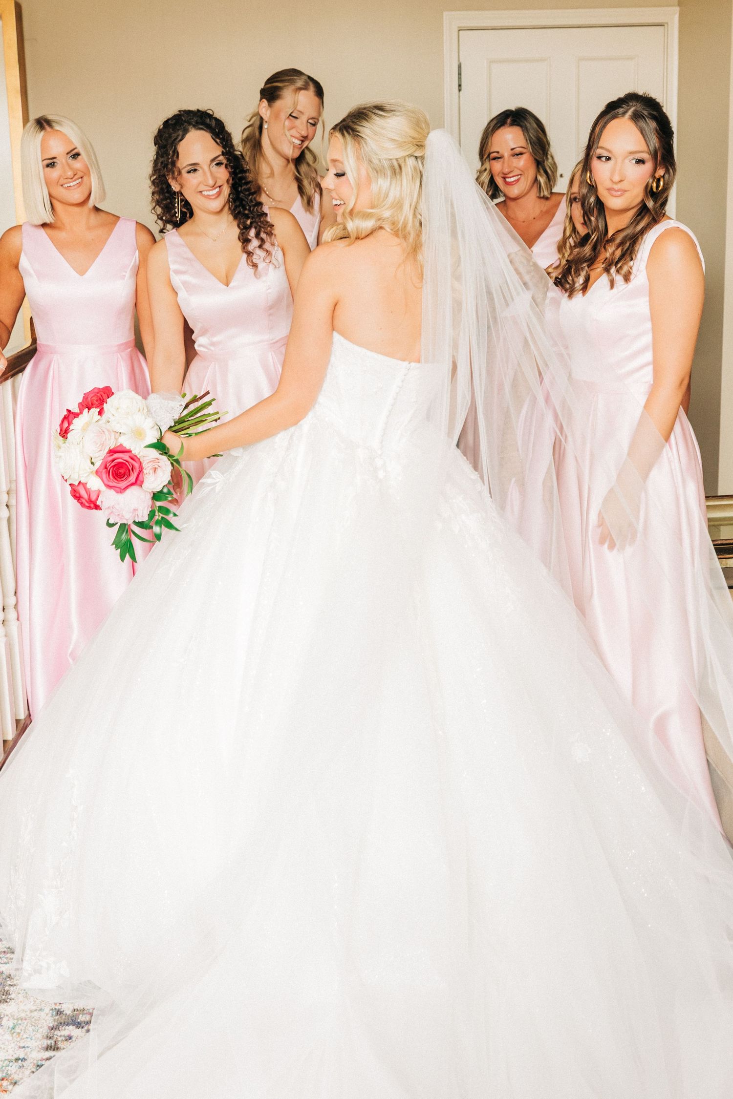 Bridesmaids in blush pink dresses gather around bride wearing white ballgown while getting ready for wedding ceremony.