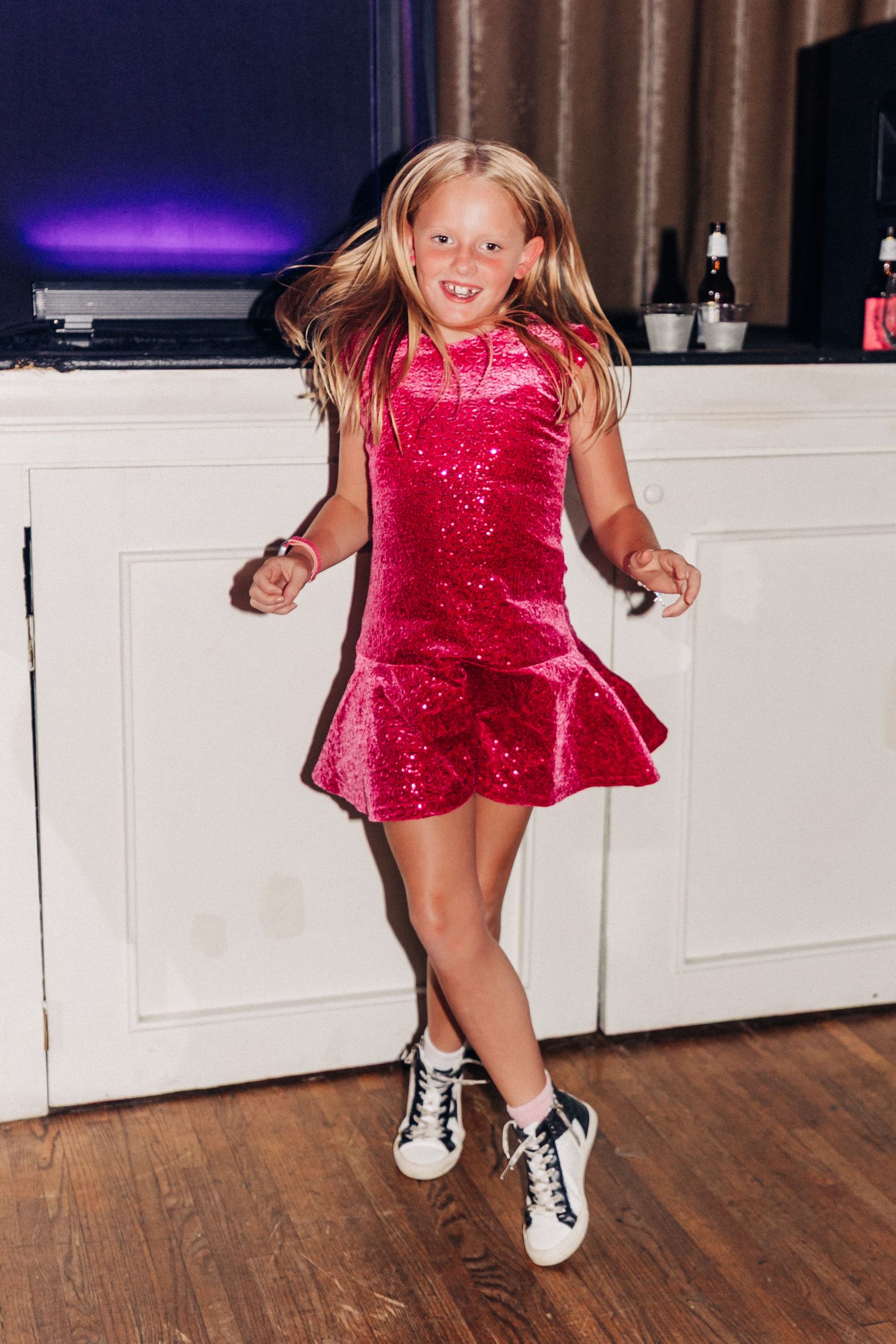 Young dancer in a sparkly red dress and sneakers jumps joyfully while dancing on a wooden floor.
