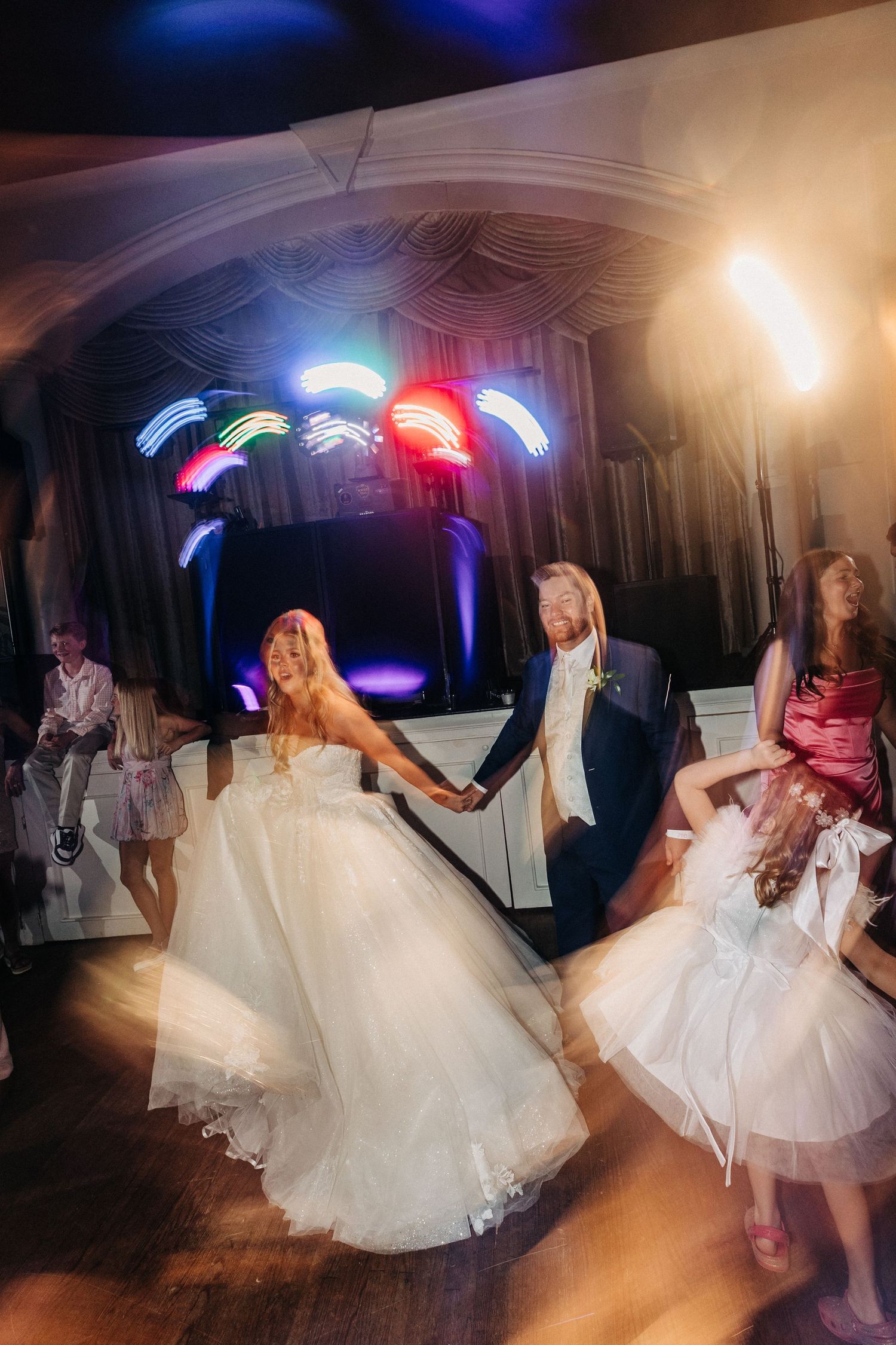 Wedding celebration on dance floor with colorful disco lights and flowing white dresses creating motion blur effect.