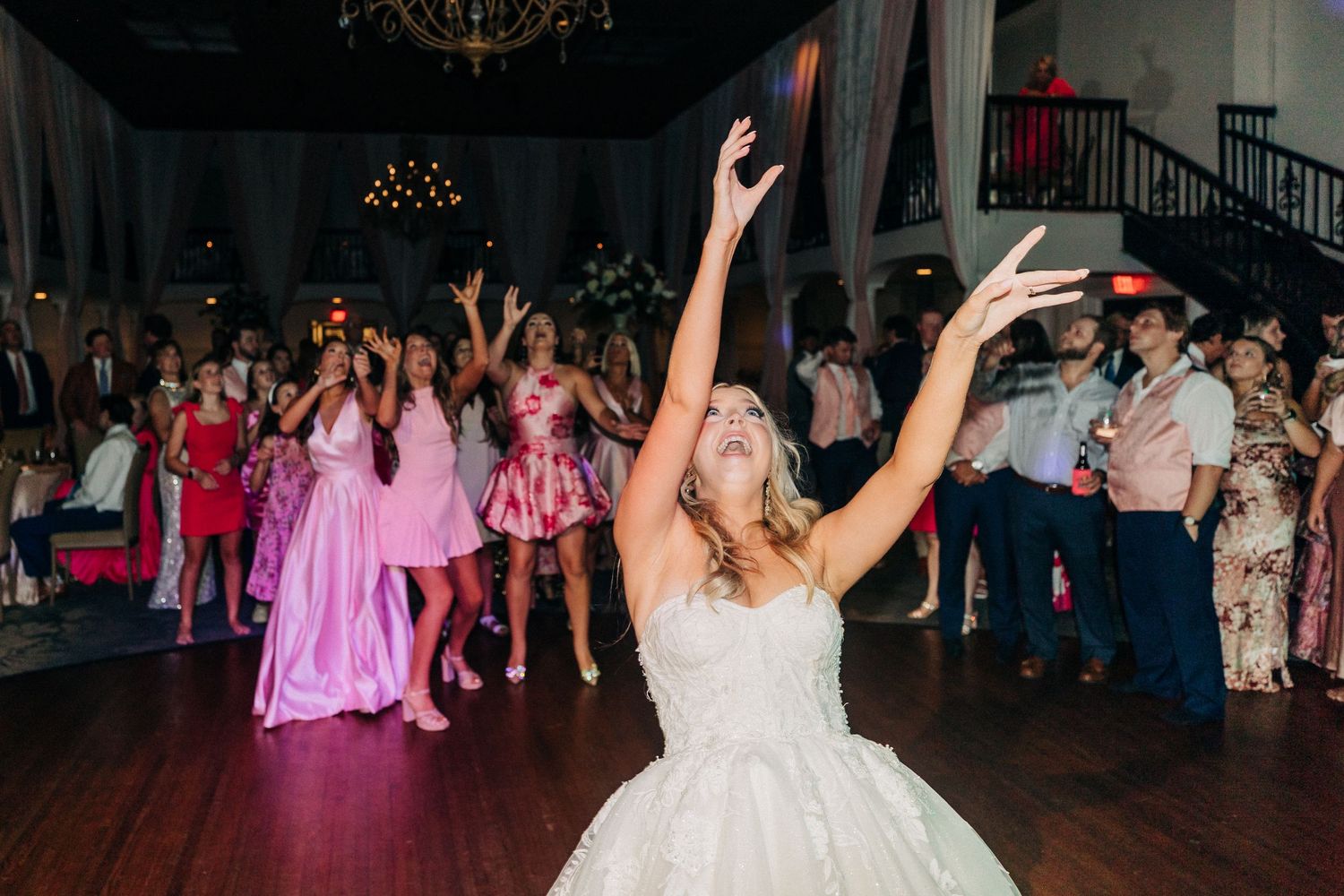 Wedding guests fill a dance floor at an evening reception with pink uplighting and a chandelier overhead.