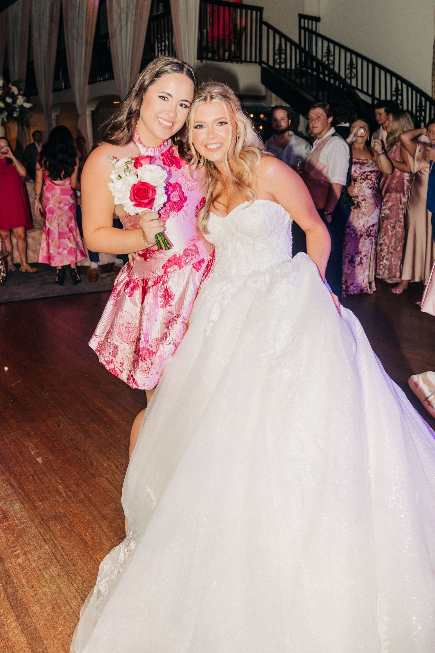 Two women pose at a wedding reception, one wearing a pink floral dress and holding a bouquet while the other wears a white gown.