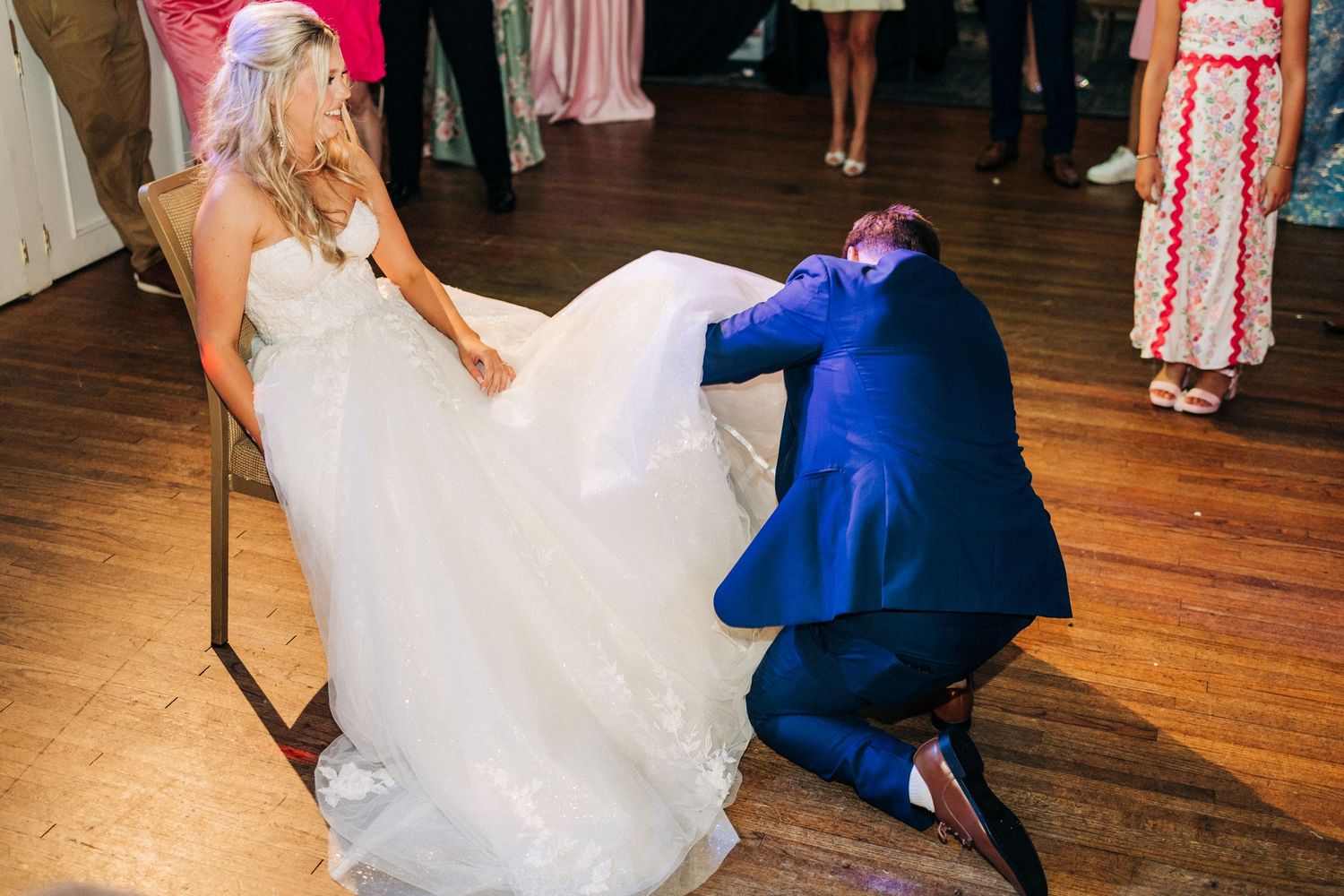 A person in a blue suit kneels to retrieve a garter from under a flowing white wedding dress on a wooden dance floor.