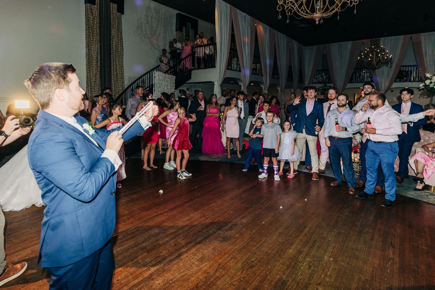 Wedding guests dance at a reception in a ballroom with a chandelier hanging overhead and rustic decor.