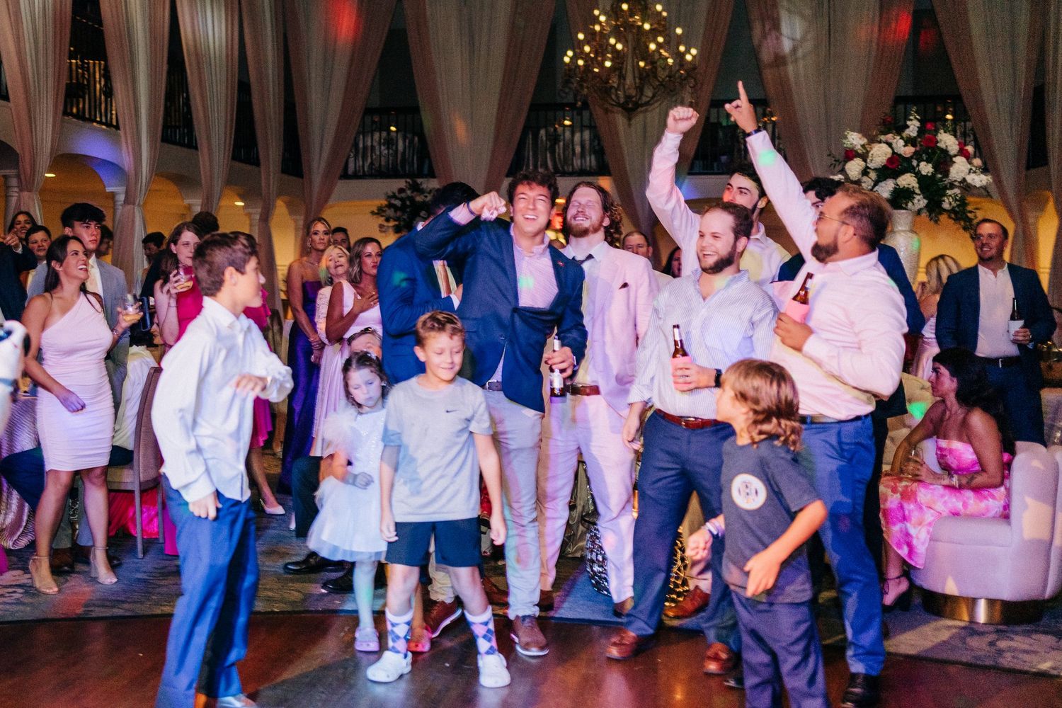 Wedding guests dance together on the illuminated dance floor during lively reception celebration with twinkle lights above.