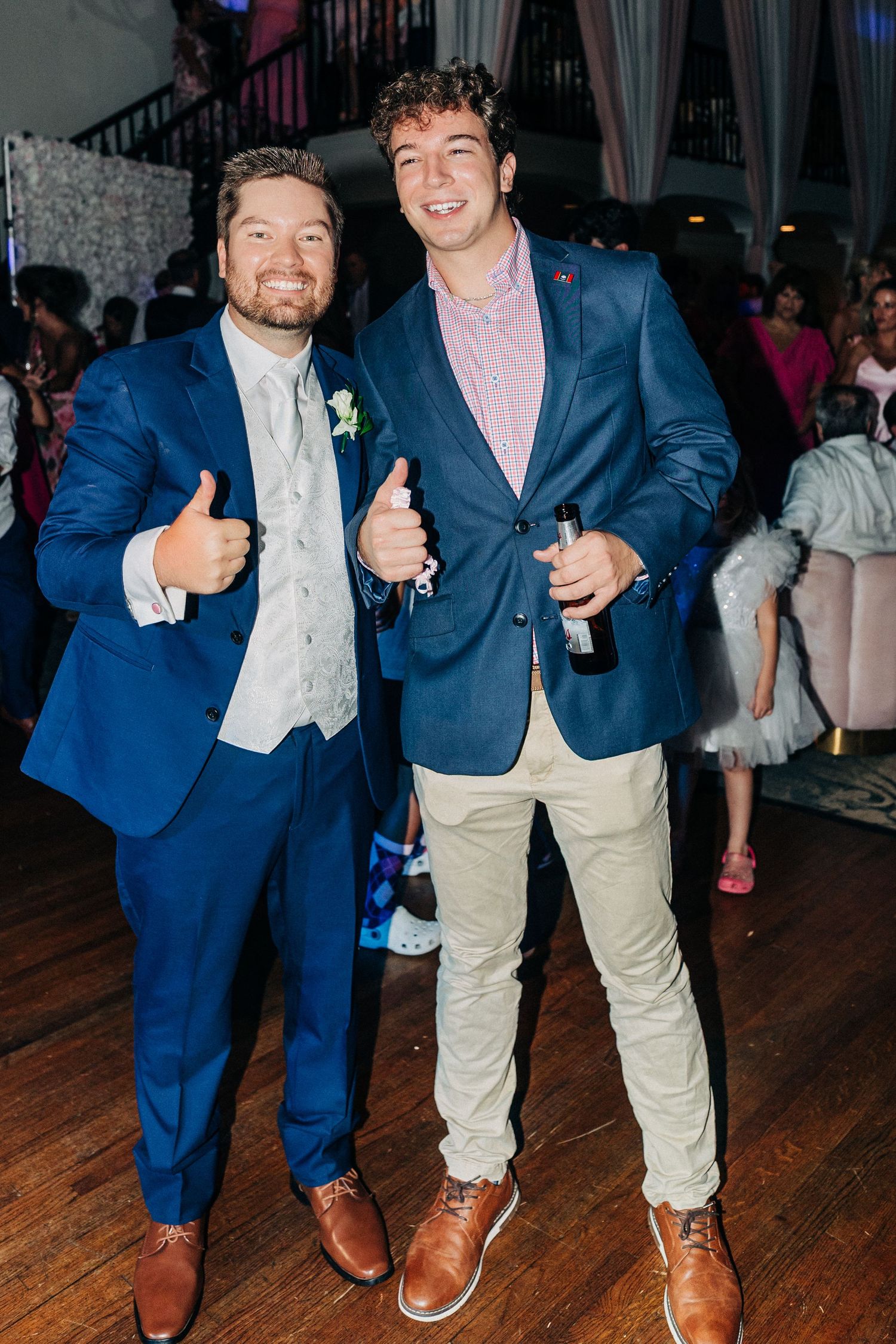 Two men in blue blazers giving thumbs up at a formal event with wooden floors and people in the background.