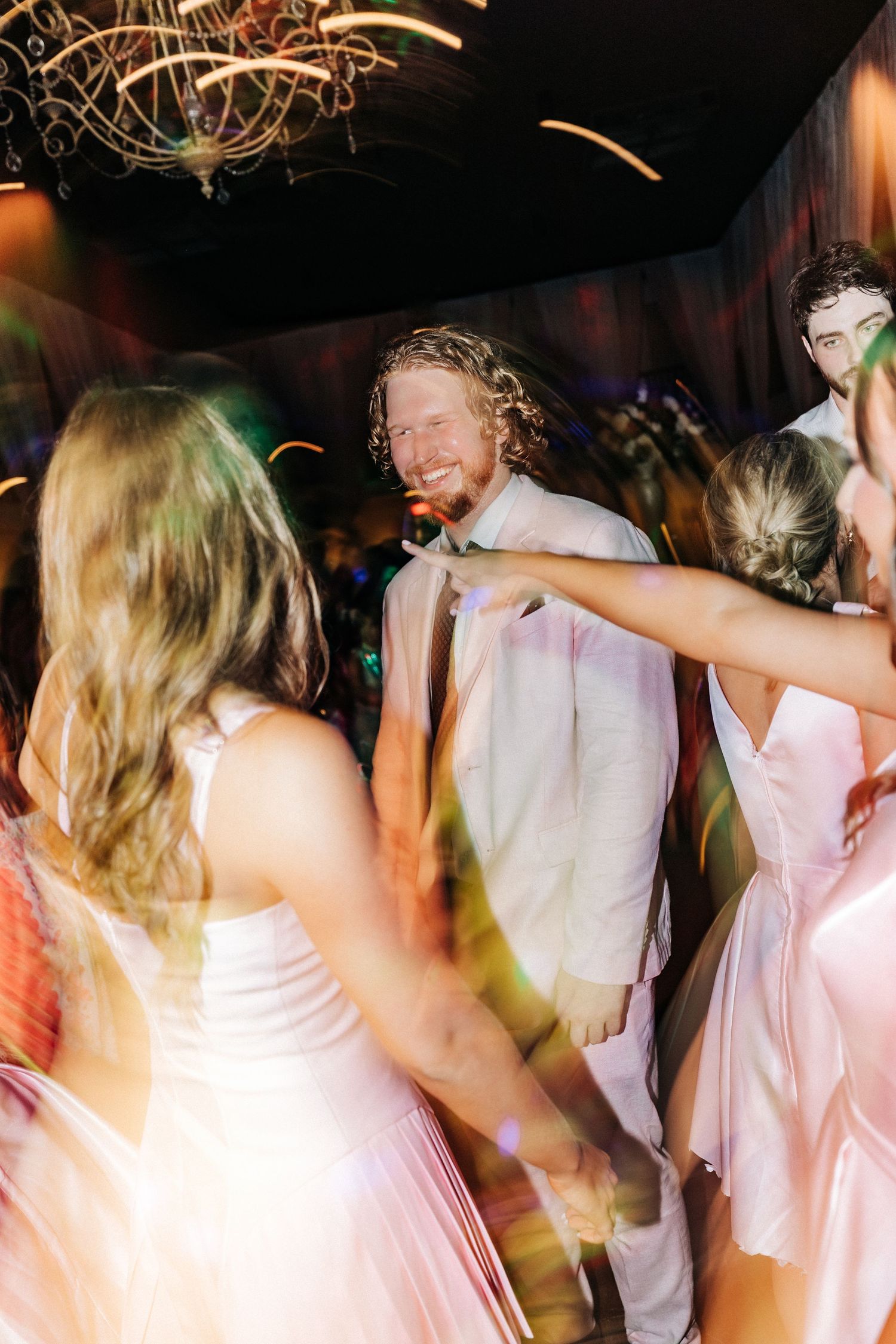 People in light colored outfits dance and celebrate at a party under a chandelier with colorful lighting effects.