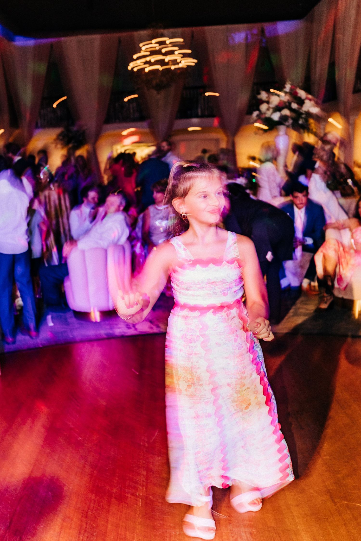 Young guest in a white dress dancing at an indoor wedding reception with purple mood lighting and a crowded dance floor.