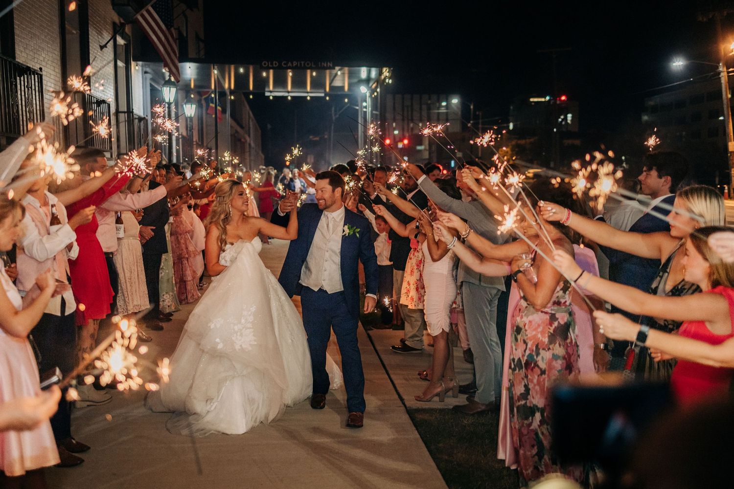 Wedding guests hold sparklers creating a magical nighttime send-off tunnel for the newlyweds during outdoor reception.