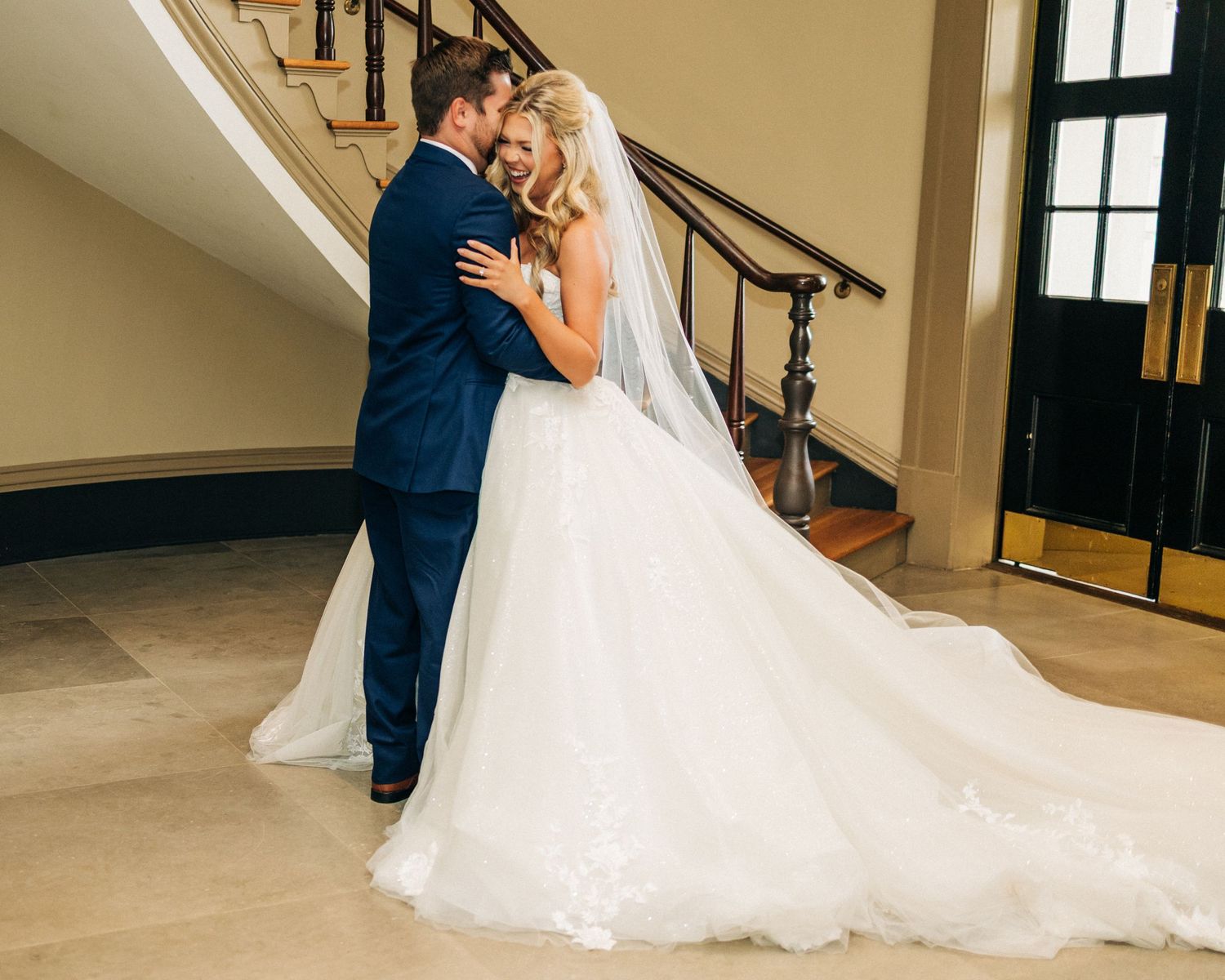 A couple shares a romantic embrace at their wedding, with the bride's flowing white gown trailing behind them near a staircase.