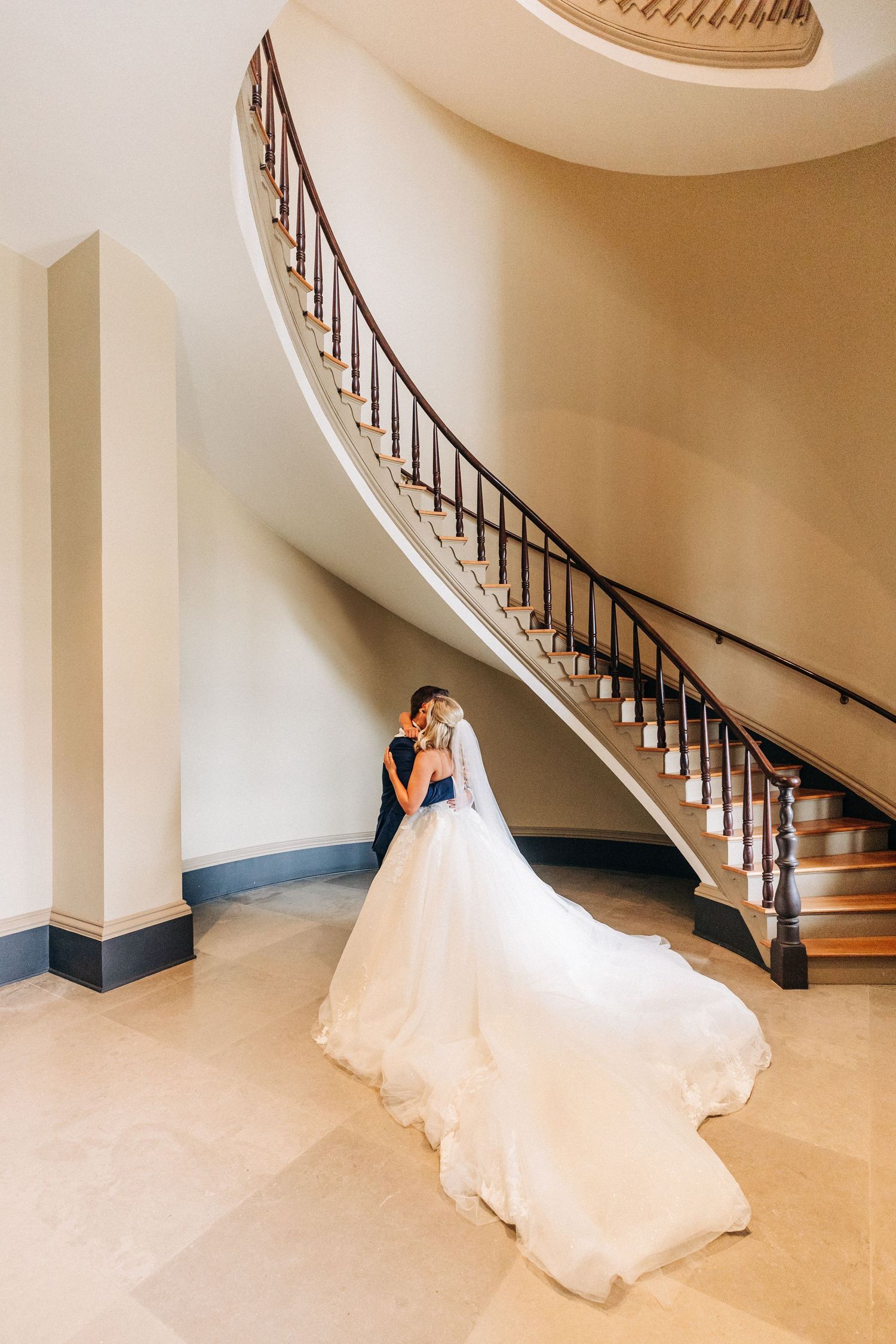 A bride in an elegant white wedding dress walks gracefully down a curved staircase with light beige walls and dark railings.