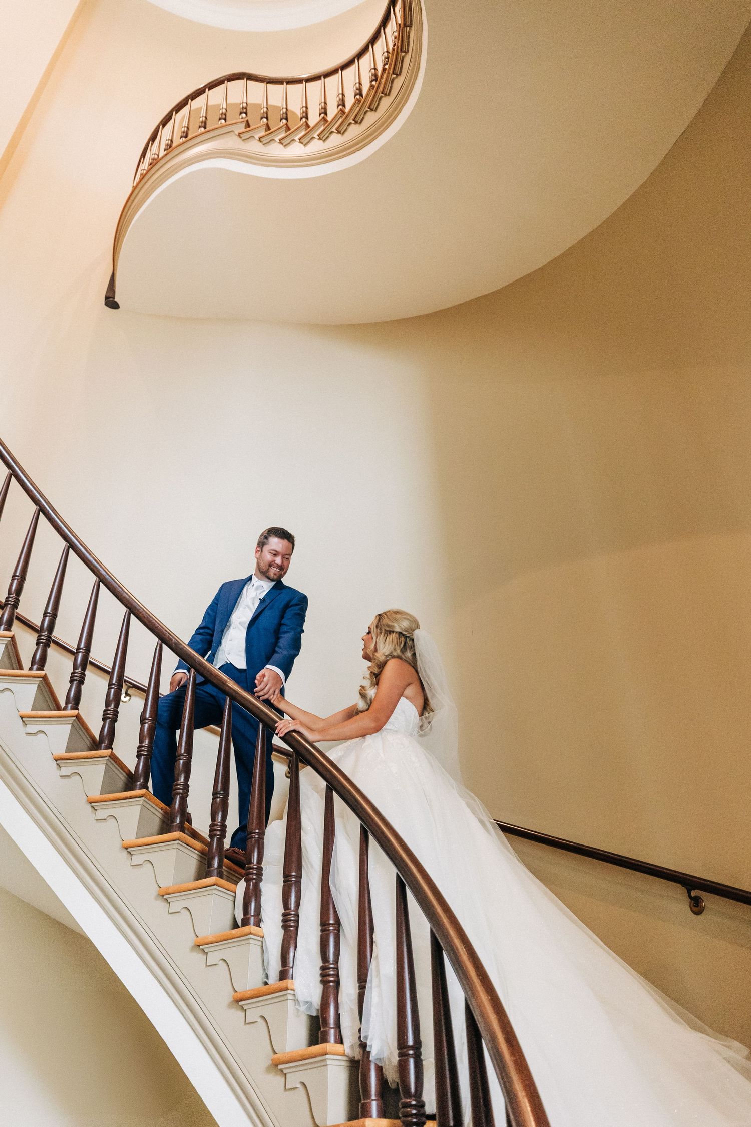 A couple poses in formalwear on a curved staircase with white walls and wooden railings during their wedding photoshoot.