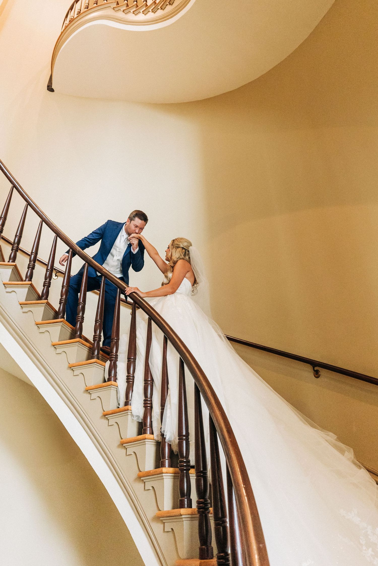 A couple stands together on an elegant curved staircase with cream colored walls and wooden banisters.