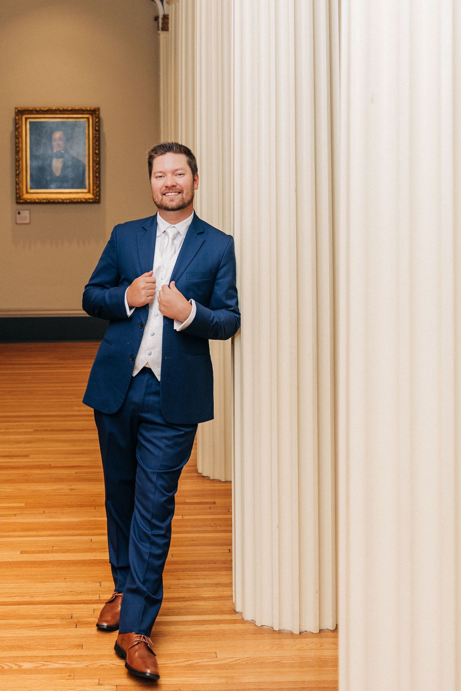 A person in a navy suit and white vest poses playfully near white columns and hardwood floors in an elegant setting.
