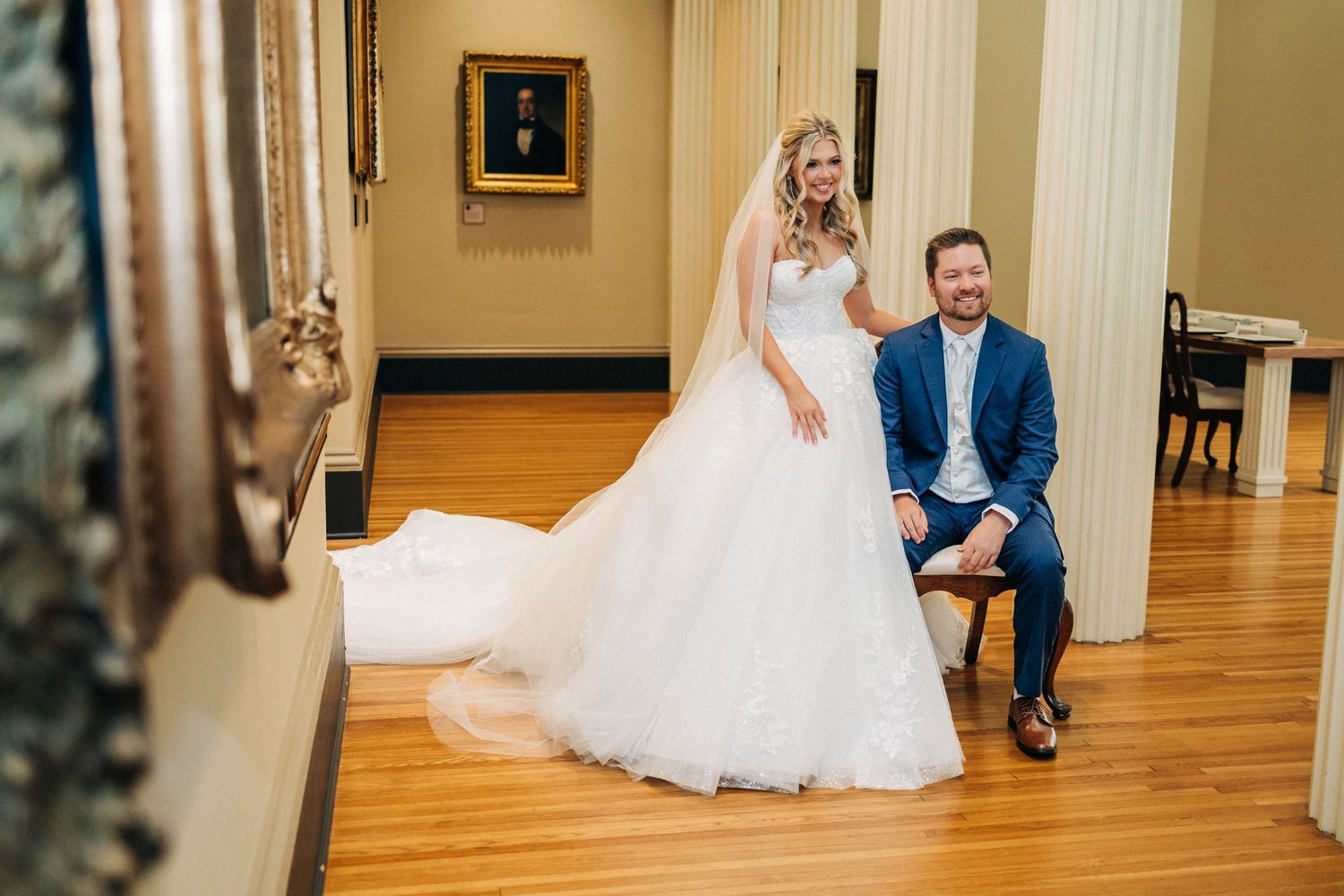 A couple in wedding attire sits together on a wooden floor in an elegant indoor hallway.