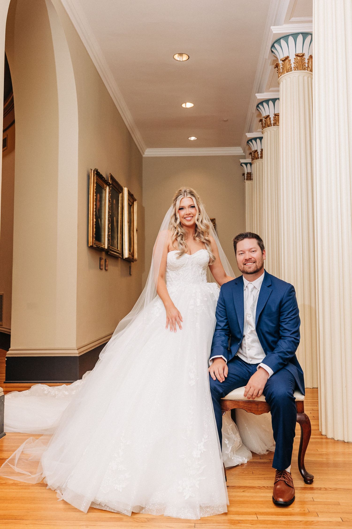 Elegant wedding scene in hallway with ornate columns and hardwood floors showing couple in bridal attire.