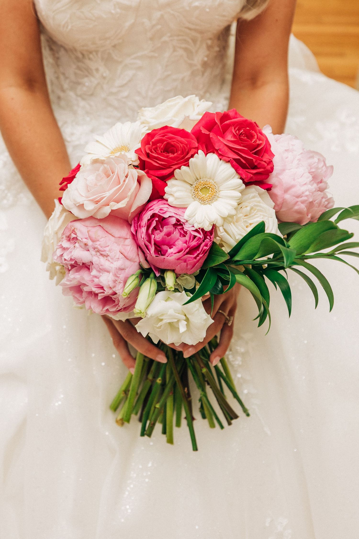 Beautiful bridal bouquet with pink and red peonies, white gerbera daisy and greenery held against white wedding dress.