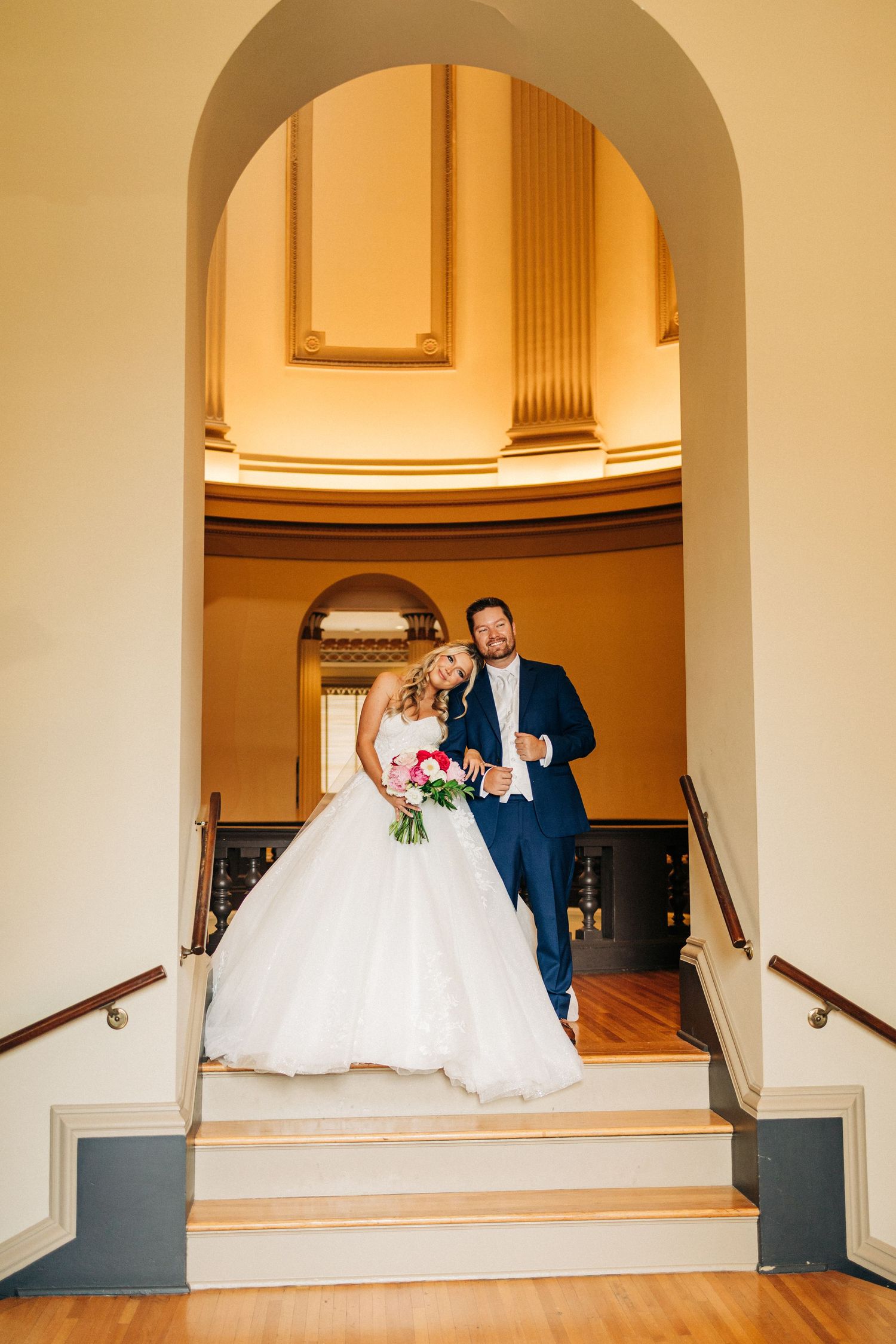 Couple in formal wedding attire dancing gracefully beneath classic arches in a warm-lit venue.