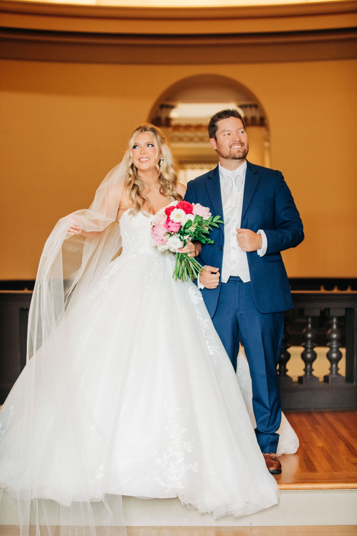 Wedding couple poses playfully together against warm indoor backdrop with elegant attire and floral bouquet.