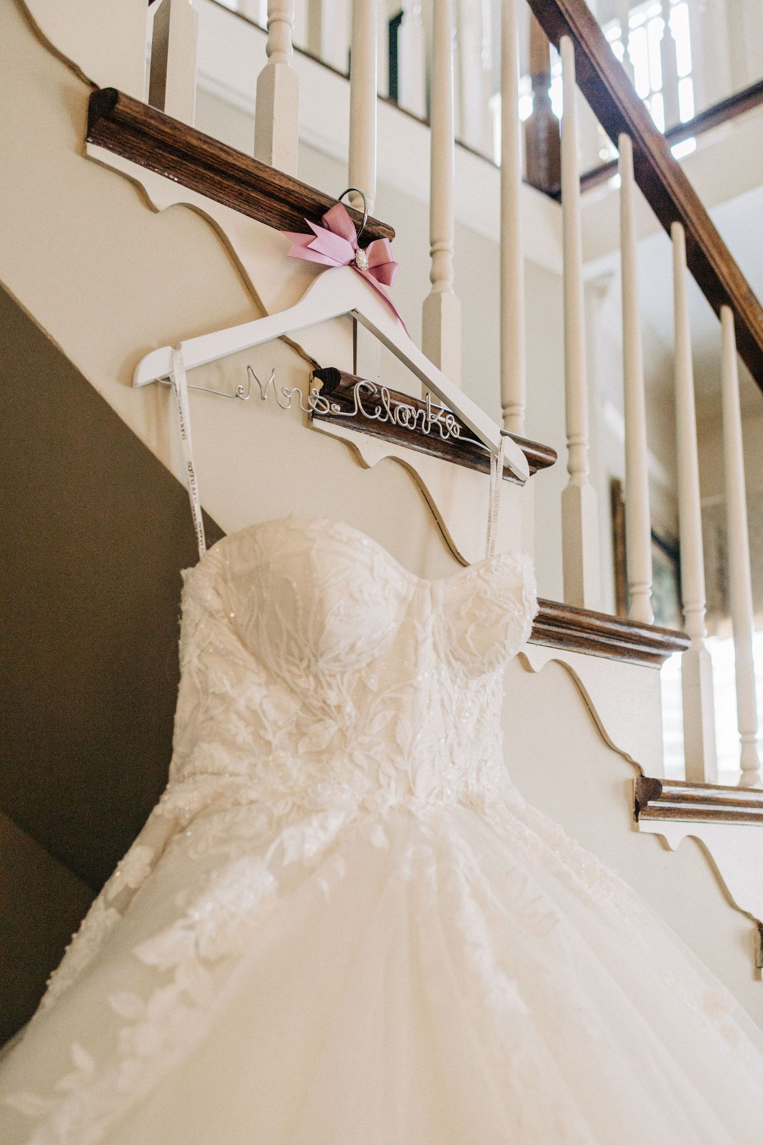 A white lace wedding dress hangs on a decorative wooden hanger against a staircase railing.