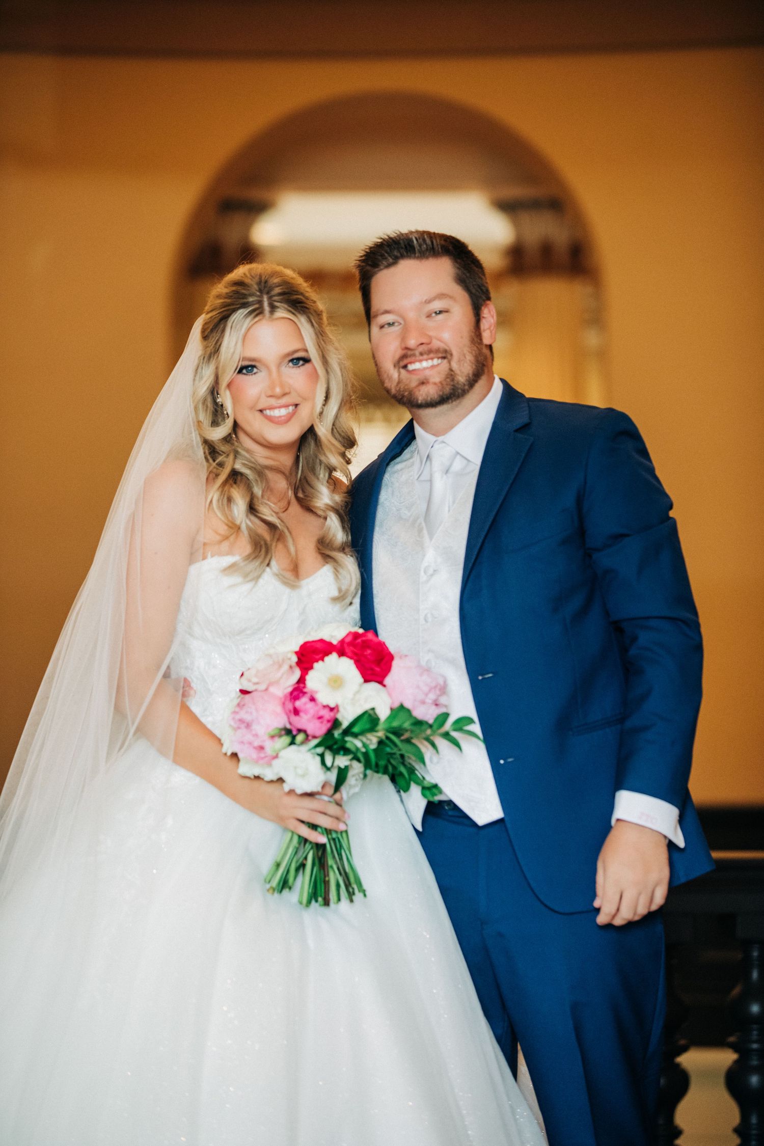 A bride holding a pink and red bouquet poses with her groom wearing a navy suit at their indoor wedding ceremony.