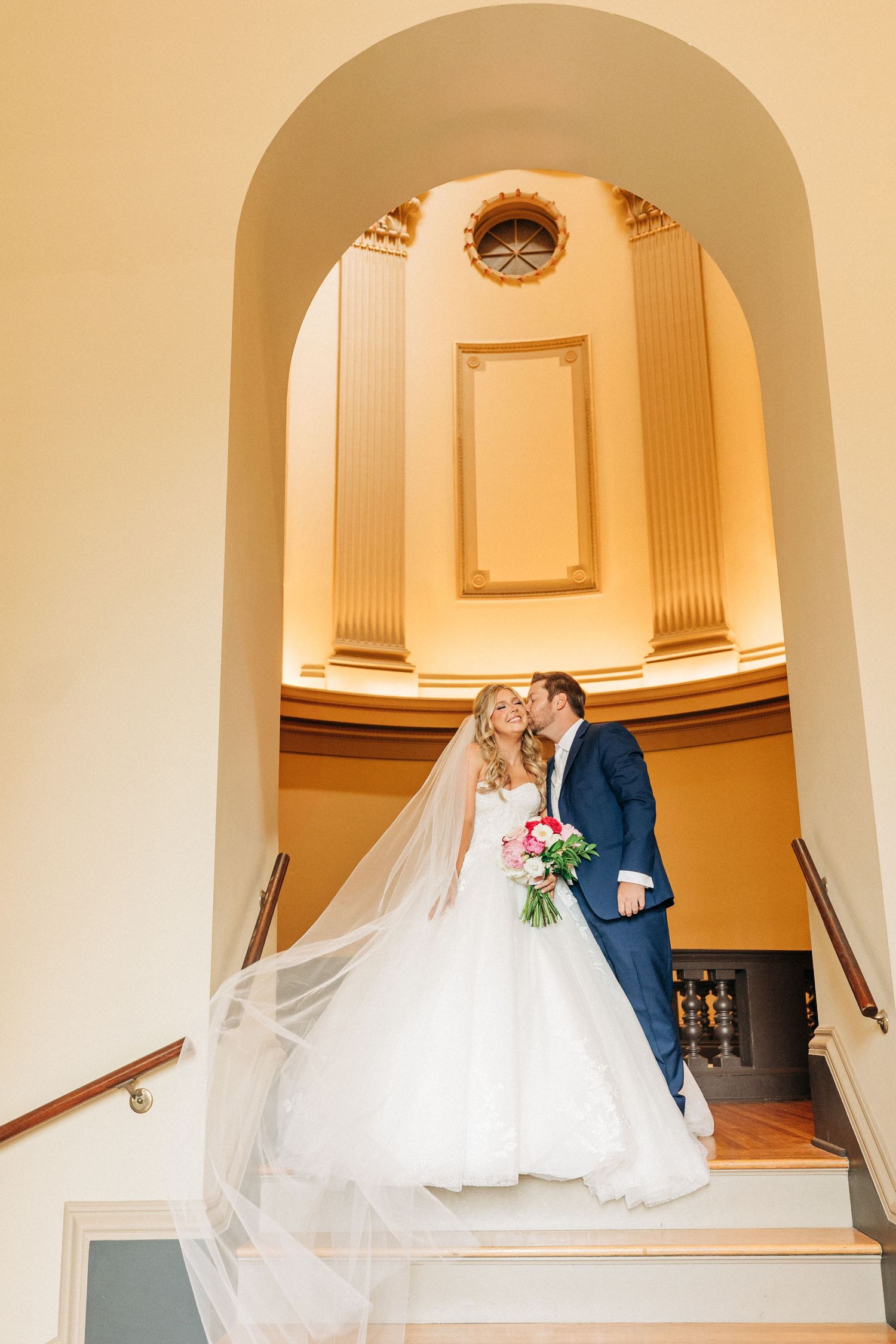 A couple in wedding attire share an intimate moment under an elegant architectural archway with cream colored pillars.